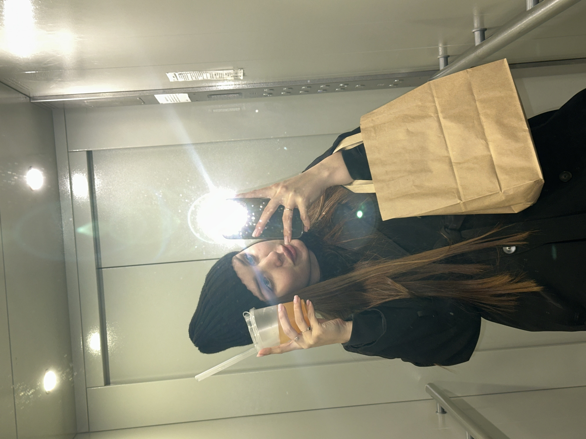 The image shows a woman taking a selfie in what appears to be an indoor, enclosed space, possibly an elevator or a modern public transport interior, in Chaikovsky, Russia. The setting features smooth, light-colored walls, overhead circular lights, and a horizontal silver bar, likely a handrail.
The woman, with long brown hair, is wearing a black beanie and a black coat. She holds a smartphone in her right hand, positioned to reflect her image, with the flash creating a bright glare on the reflective surface. In her left hand, she holds a clear cup containing a light-colored beverage with a white straw. Two silver rings are visible on her fingers. Behind her left shoulder, a brown paper bag with handles is visible, hanging or being carried. Her expression is calm as she looks towards the camera's reflection.
Notable details include the bright artificial lighting and the modern, minimalist design of the interior space. Small, unreadable text or markings are visible on a metallic strip near the ceiling in the background. The time of day and weather are not discernible due to the indoor setting.