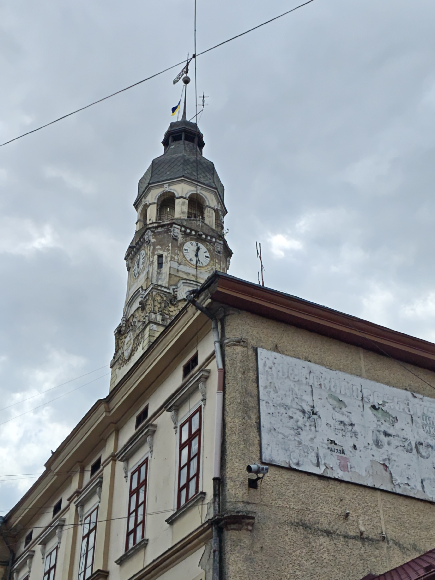 The image presents a low-angle view of an old clock tower and a section of an adjacent building in Sniatyn, Ukraine, under a heavily overcast sky. The prominent clock tower features weathered stone or plaster construction with ornate classical architectural elements, including decorative carvings, columns, and arched openings leading to a belfry. At least two clock faces are visible, both indicating approximately 12:05 or 12:55. The tower is crowned with a dark, domed roof and a spire, from which a weather vane and a small Ukrainian flag (blue and yellow) are proudly displayed. Utility cables diagonally traverse the grey sky in front of the tower.
To the right, part of an adjoining building is visible, characterized by its textured, somewhat worn walls, possibly stucco or concrete. A large, rectangular billboard or sign covers a significant portion of this wall, but it is heavily defaced and peeling, rendering most of its printed content illegible, although faint Cyrillic characters can be discerned. A security camera is mounted on the wall below the billboard. The building's windows feature striking reddish frames with multiple panes, encased in white decorative surrounds. A brown fascia board and gutter system mark the roofline. The scene depicts historical urban architecture amidst a cloudy daytime environment, showcasing signs of age and everyday urban elements.