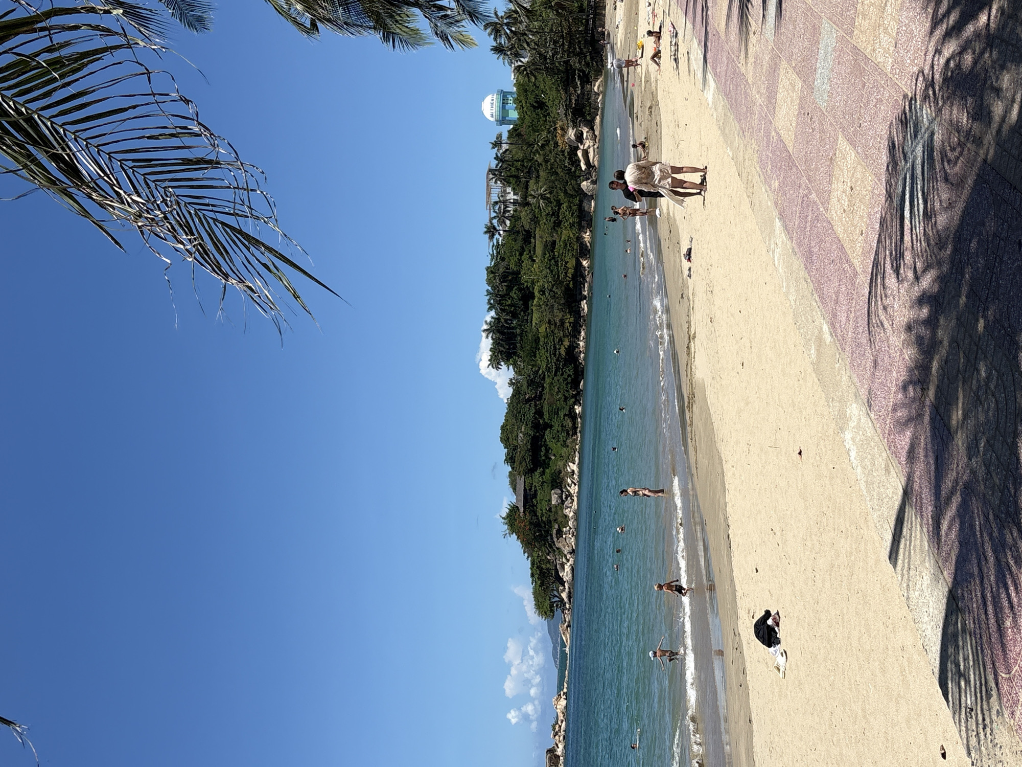 The photograph captures a beach scene in Bac Nha Trang Ward, Vietnam, under a clear blue sky. The beach is lined with a paved walkway, patterned in pink and tan squares, partially obscured by the shadow of a palm tree. The sandy beach leads to turquoise water, where several people are swimming and wading. On the beach, a few individuals are walking or standing near the shoreline, while others are positioned further up the beach. Lush, green vegetation covers a hill behind the beach, with a tall, cylindrical water tower visible among the trees. A couple stands together on the beach. In the upper left corner, the fronds of a palm tree frame the sky.