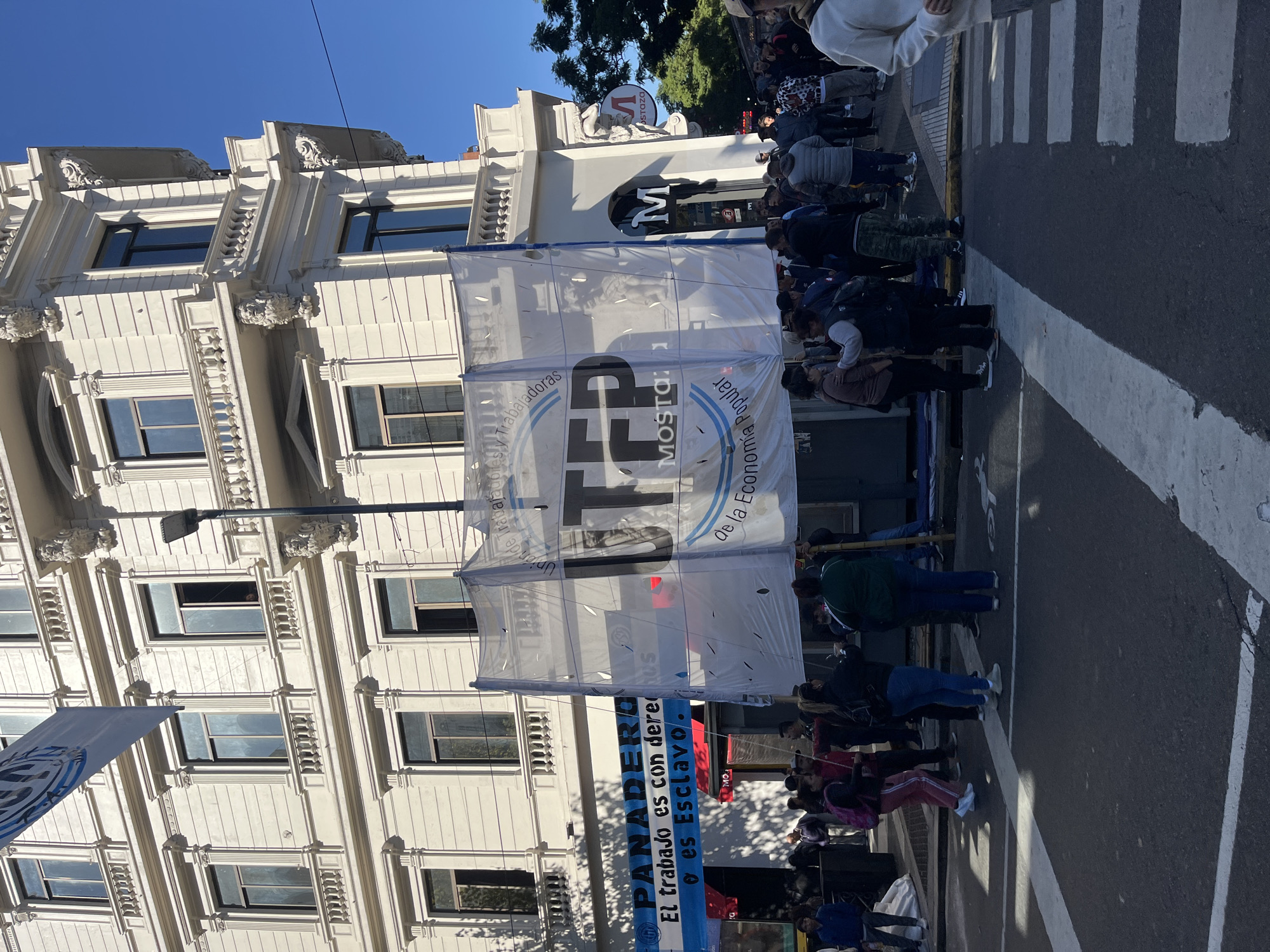 The image depicts a daytime street scene in Buenos Aires, Argentina, showing a public gathering or demonstration. Numerous people are present, congregating on and alongside the street. The sky is clear and bright, indicating sunny weather.
Prominently displayed in the center is a large, white translucent banner stretched across the street. Visible text on this banner includes "Unión de Trabajadores y Trabajadoras" (Union of Workers - male and female), "FP", "MOSTO: 1", and "de la Economía Popular" (of the Popular Economy), suggesting the presence of a workers' union, likely the FENAT Mosto branch of UTEP (Union of Workers of the Popular Economy). Below this, a blue banner is partially visible, bearing the word "PANADERO" (Baker) and the slogans "El trabajo es con derechos" (Work is with rights) and "es Esclavo." (is Slavery), advocating for fair labor practices.
In the background, an ornate, light-colored building with multiple windows and decorative architectural details, such as cornices and pilasters, dominates the left side of the frame. Further down the street, another building features a sign with a large "M" and "STAZA" or "MOSTOZO". A pedestrian crosswalk with white stripes is visible on the asphalt. The scene suggests a protest or public rally focused on workers' rights.