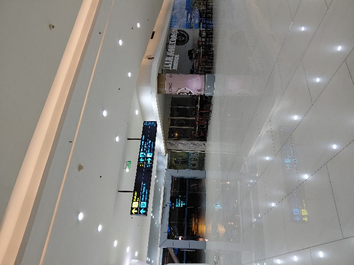 A bright, expansive indoor space showcases a modern building interior. White tile flooring reflects overhead lighting. Signage directs travelers to various destinations, including parking and gates. Multiple restaurants and shops line the upper level. The overall impression is clean and spacious, evoking a contemporary airport or shopping mall.