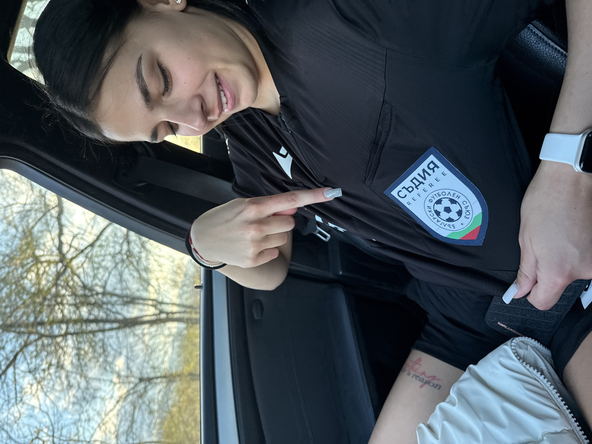 A woman is in a vehicle. She is wearing a black referee jersey with a patch that says "СЪДИЯ REFEREE" and includes the Bulgarian flag. She is pointing at the patch with her right index finger. She is wearing a white watch on her right wrist. Part of a tattoo is visible on her thigh. The view out of the vehicle window is blurry, but shows trees and the sky. The location is not apparent from the image. There is no information to suggest this photo was taken in Gorna Rositsa, Bulgaria.