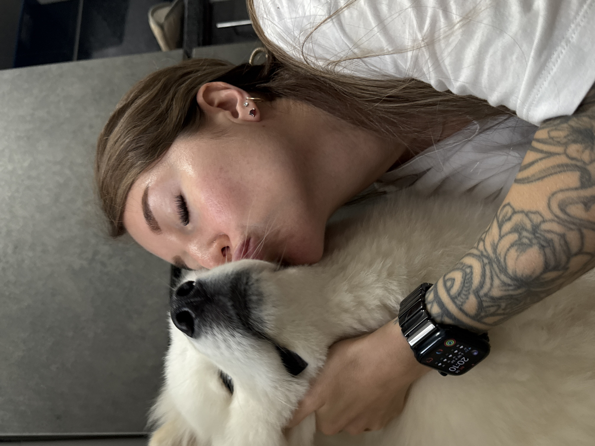 A woman with brown hair and visible tattoos on her arm is kissing a white fluffy dog. She is wearing a white t-shirt and a black smartwatch. The dog's nose and snout are visible. The background appears to be an indoor setting with a grey textured wall and some shelving units. The image does not contain information about the city of Chaikovsky, Russia.