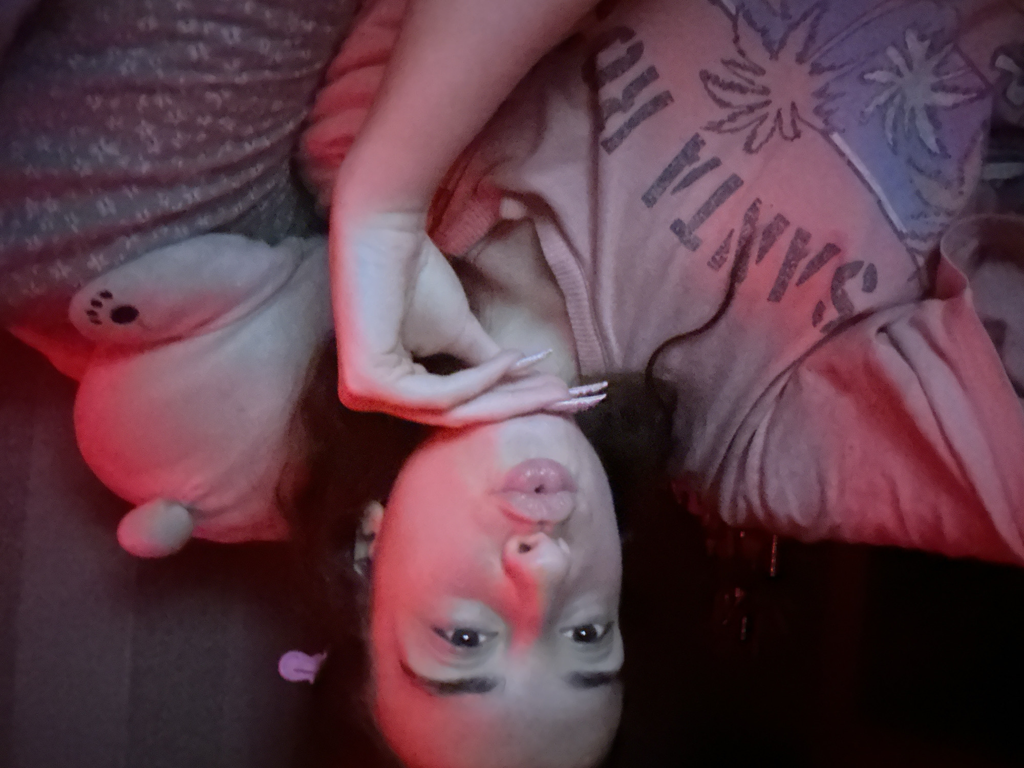 A young woman is captured in a close-up selfie, her face illuminated by red light, which casts a warm glow on her skin. She is puckering her lips, giving a kissy face, and her right hand is gently resting on her chin. Her long, manicured fingernails are visible, adorned with subtle white detailing. She's wearing a light-colored t-shirt with a graphic design that includes the word "SANTA" prominently displayed, along with palm tree motifs. To her right, a plush toy with a small, round ear and a paw print detail is visible, partially obscured by her body. The background appears to be a dimly lit indoor space, possibly a bedroom, with a dark wall and what looks like textured bedding. The overall impression is of a casual, personal moment, likely taken at home. There is no discernible information to suggest a location in Shumen, Bulgaria.