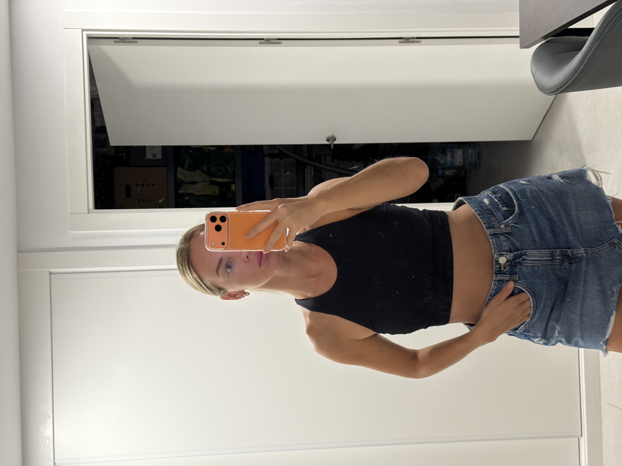 The image is a mirror selfie taken by a woman with blonde hair in a black crop top and a denim skirt. She is holding an orange smartphone in her right hand, which is obscuring her face. Her left hand is in her skirt pocket. The background shows a white door and a partially open closet door revealing shelves with various items. The flooring is light-colored. The scene appears to be an indoor residential setting, likely a closet or hallway. No specific details are present to indicate the location is in Finestrat, Spain.