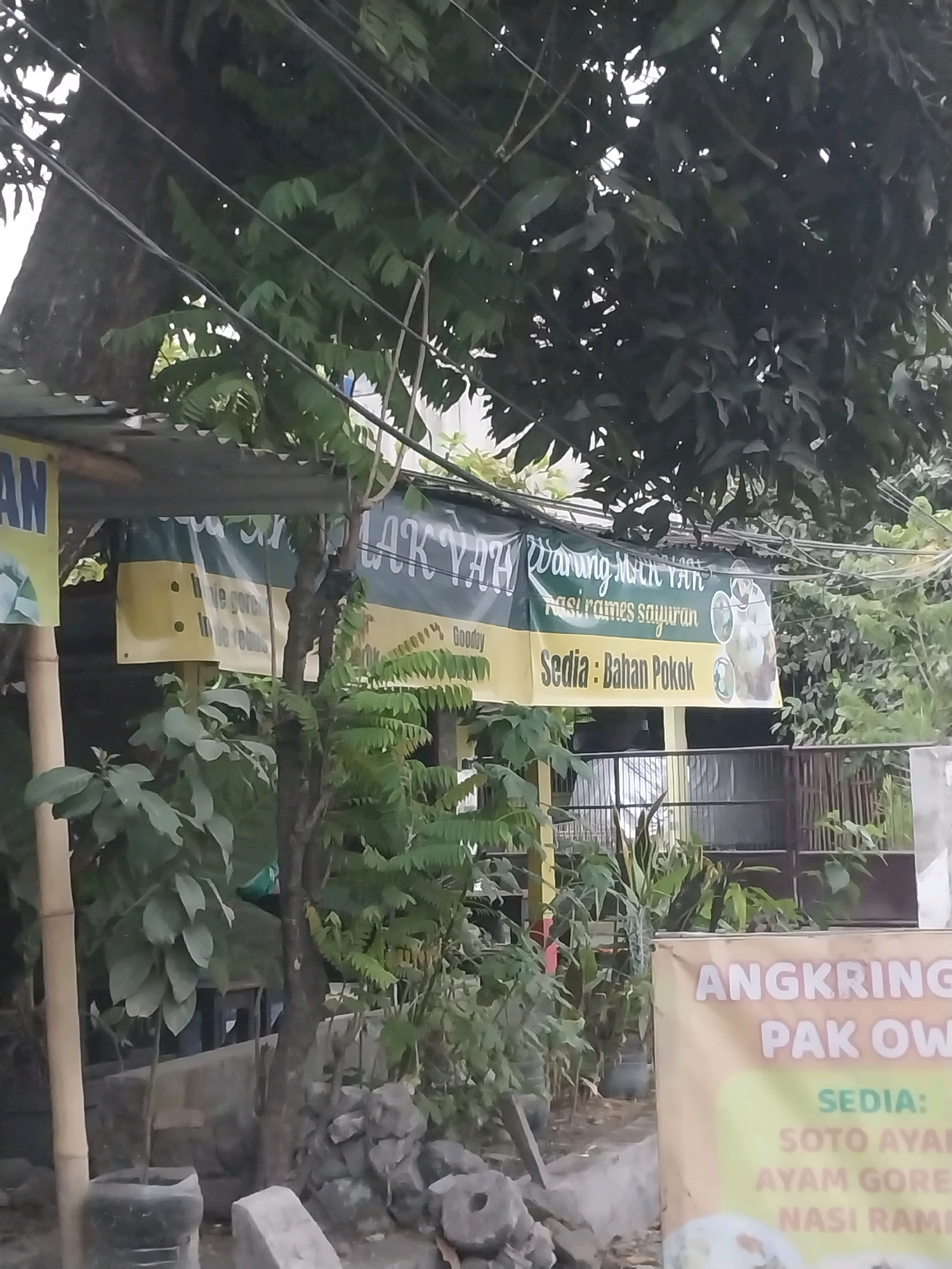 The image shows a street-level view of a small food stall in Maguwoharjo, Indonesia. A green and yellow banner hangs across the front of the stall, displaying text in Indonesian, including "Warung Mak Yah," "Nasi rames sayuran," and "Sedia: Bahan Pokok." To the right of the banner, another sign is partially visible with the text "ANGKRING," "PAK OW," and "SEDIA: SOTO AYAM, AYAM GORENG, NASI RAM."
The stall is situated outdoors, with a corrugated metal roof supported by a wooden structure. Lush green foliage from trees and plants surrounds the stall, creating a natural and somewhat overgrown environment. Some of the plants appear to be potted, while others grow directly from the ground. In the background, a fence and some buildings are visible, suggesting an urban or semi-urban setting. There are no people clearly visible in the image, but the presence of the food stall indicates commercial activity. The overall scene suggests a simple, local establishment offering food and basic provisions. The lighting suggests it is daytime.