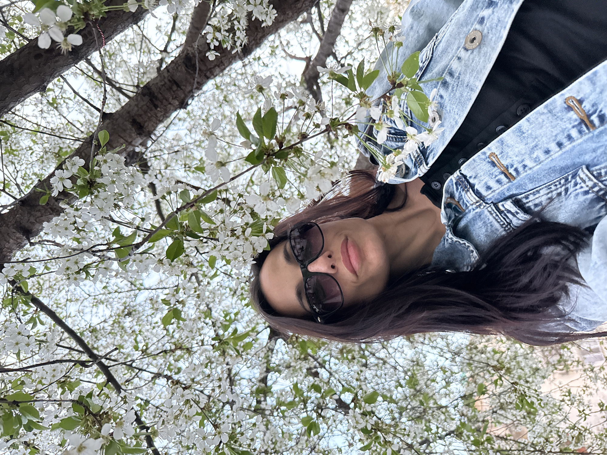 A woman with sunglasses and a denim jacket is taking a selfie under a tree with white blossoms. The location is Pleven, Bulgaria.