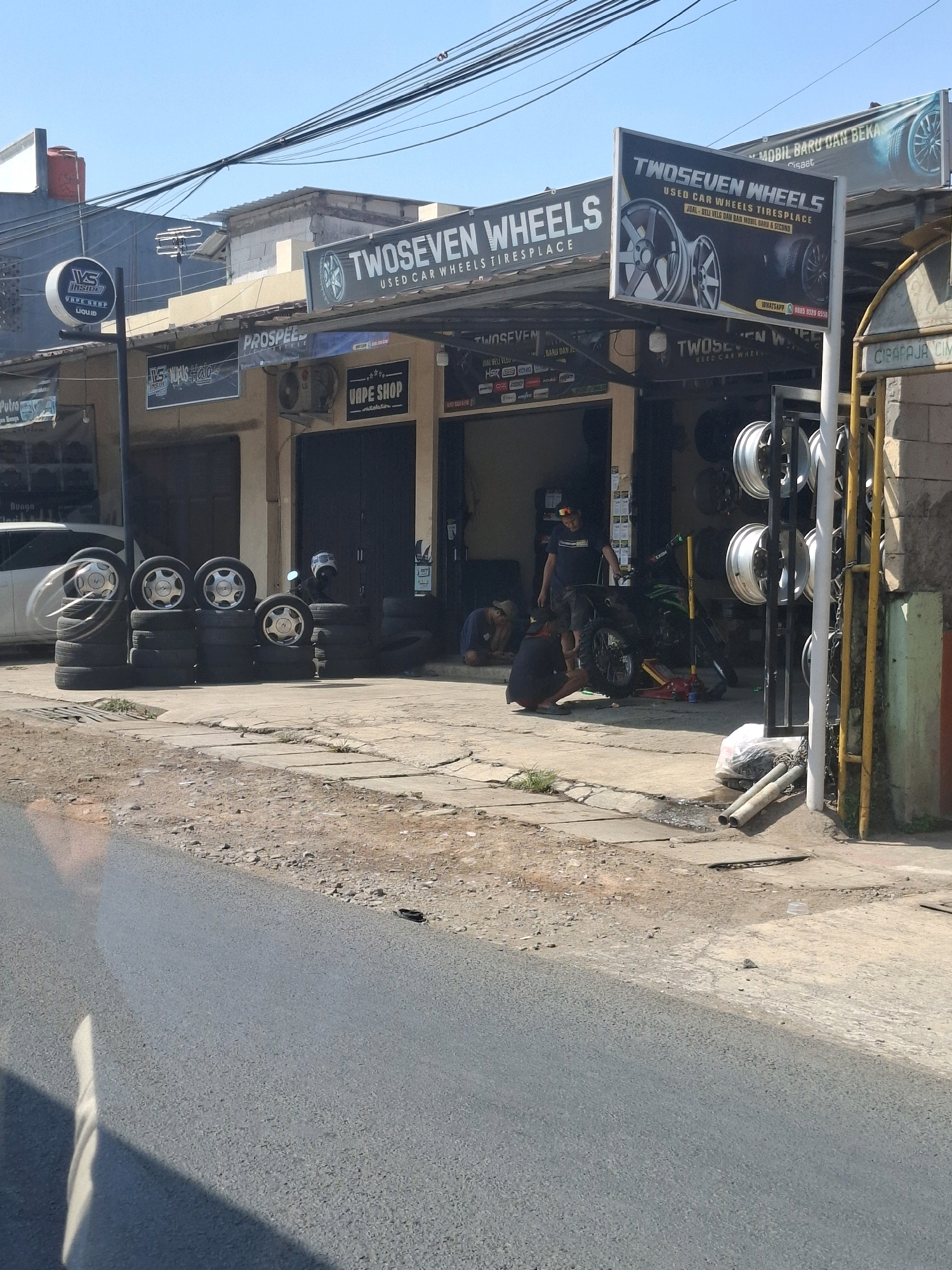 The image depicts an outdoor scene in Selajambe, Indonesia, likely a commercial area. In the foreground, a paved road runs diagonally across the lower portion of the frame, with a dirt and gravel shoulder to its right. The main focus is a car wheel and tire shop. A large sign above the shop reads "TWOSEVEN WHEELS USED CAR WHEELS TIRESPLACE" with additional text indicating it is a "JUAL BELI VELG & BAN MOBIL BARU & SECOND" (buying and selling new and used car rims and tires). To the left of this sign, another sign reads "PROSPEED" with smaller text underneath.
Several stacks of used car tires are arranged on the sidewalk in front of the establishment. Inside the open-front shop, two individuals appear to be working on a motorcycle. One person is kneeling next to the motorcycle, and another is standing nearby. A green motorcycle is present, along with a red hydraulic jack. A display rack of several car rims is visible to the right of the shop entrance. The building itself has a dark facade with shop windows. Above the shop, overhead power lines are visible against a clear blue sky. The environment appears to be sunny with shadows cast on the ground.