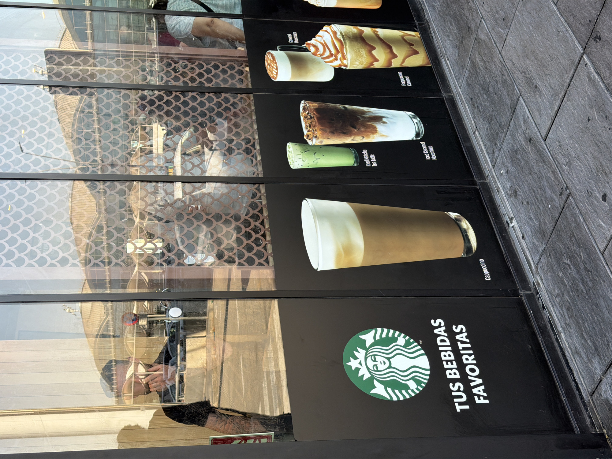 A man in a black t-shirt is reflected in a glass window with a patterned design. He is holding a phone to his ear. Below the reflection, a black panel displays the Starbucks logo and the text "TUS BEBIDAS FAVORITAS" in white lettering. To the right of the logo, there are images of four drinks: a cappuccino, an iced matcha tea latte, an iced caramel macchiato, and a caramel Frappuccino. The ground in front of the window is paved with gray stone tiles. The scene appears to be in Elx/Elche, Spain, as indicated by the Spanish text on the sign.