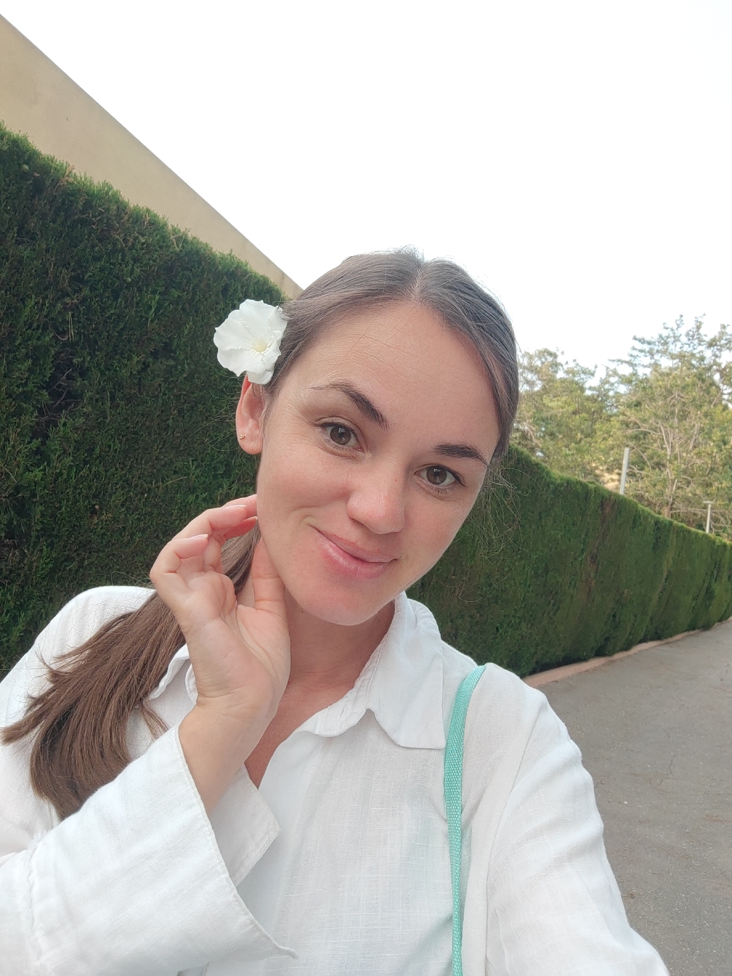 A selfie taken outdoors in El Campello, Spain, shows a woman with a white flower behind her right ear. She is wearing a white linen shirt with a light blue strap across her shoulder. The woman is looking at the camera and smiling, with her right hand touching her neck. Behind her, a well-maintained green hedge runs along a paved walkway, with trees visible in the distance. The sky above is a bright white, indicating daylight. The scene suggests a pleasant, possibly warm, day.