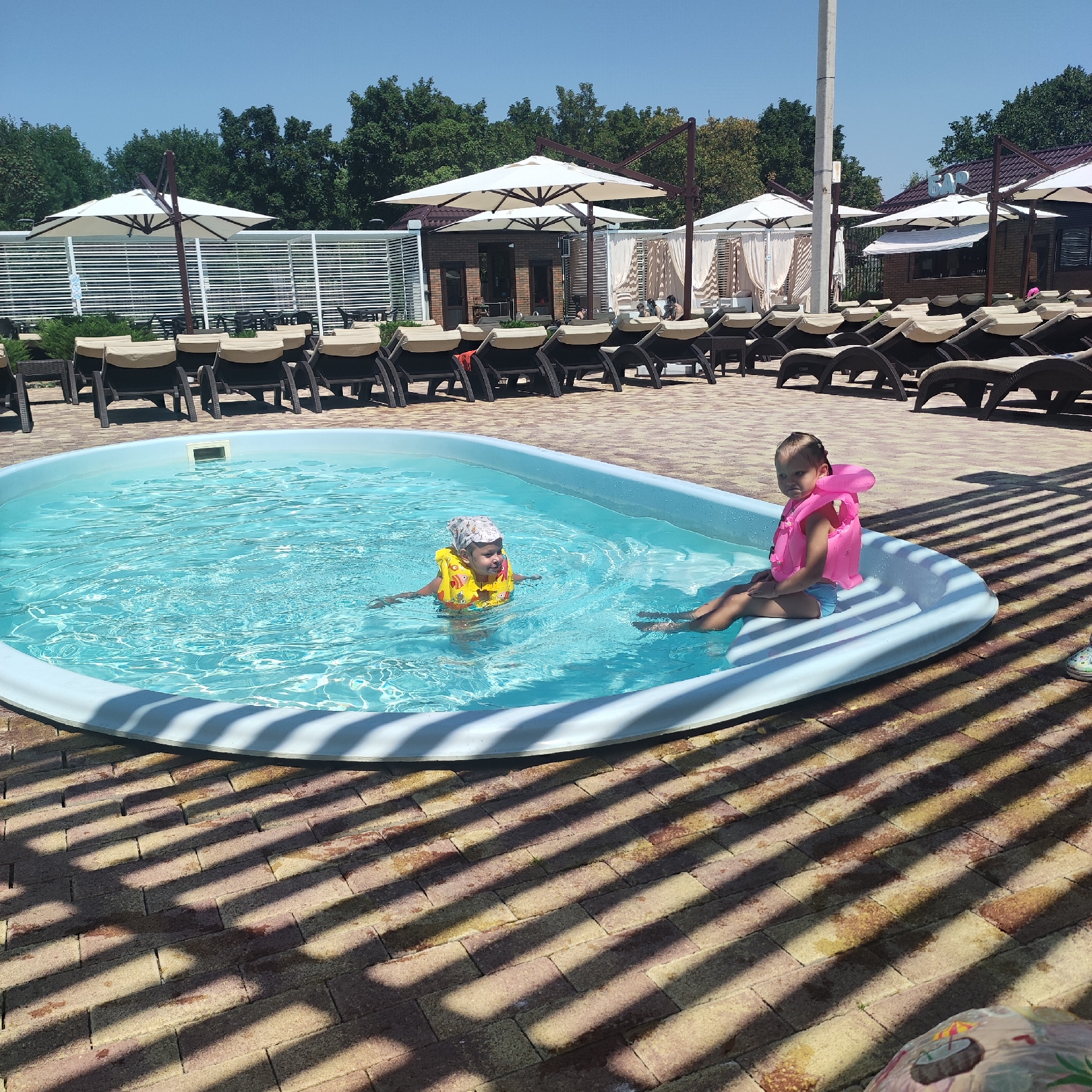 In an outdoor setting with a blue sky and lush green trees in the background, a kidney-shaped swimming pool is visible. The pool is filled with clear blue water, and two young children are present. One child, wearing a yellow life vest and a bandana, is in the water, appearing to swim. Another child, wearing a pink life vest and blue shorts, is seated on the edge of the pool, facing the camera. The pool area is paved with brick-like tiles, and there are lounge chairs and large white umbrellas set up in the background, suggesting a resort or recreational area. Shadows from a slatted structure are cast across the tiled ground.