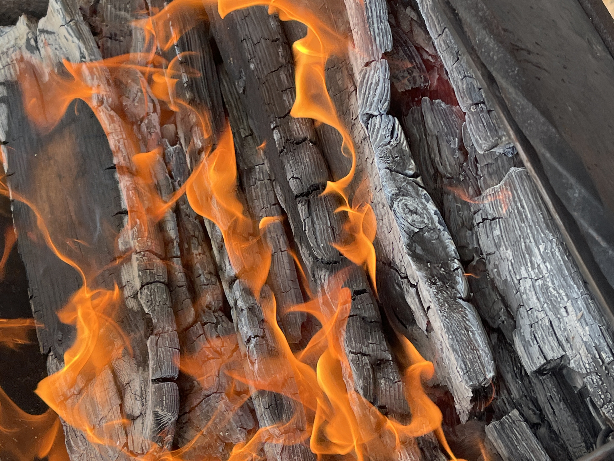 The image is a close-up, vertically oriented shot of burning logs in Il'ino, Russia. The logs are dark gray and charred, with prominent cracks and grain patterns visible. Bright orange and yellow flames dance and flicker across the surfaces of the wood, obscuring some areas and highlighting the texture of the burning material. The background is dark and out of focus, emphasizing the fire and wood. There are no people or animals visible in the image. The scene appears to be an outdoor setting, possibly a bonfire or a fireplace.