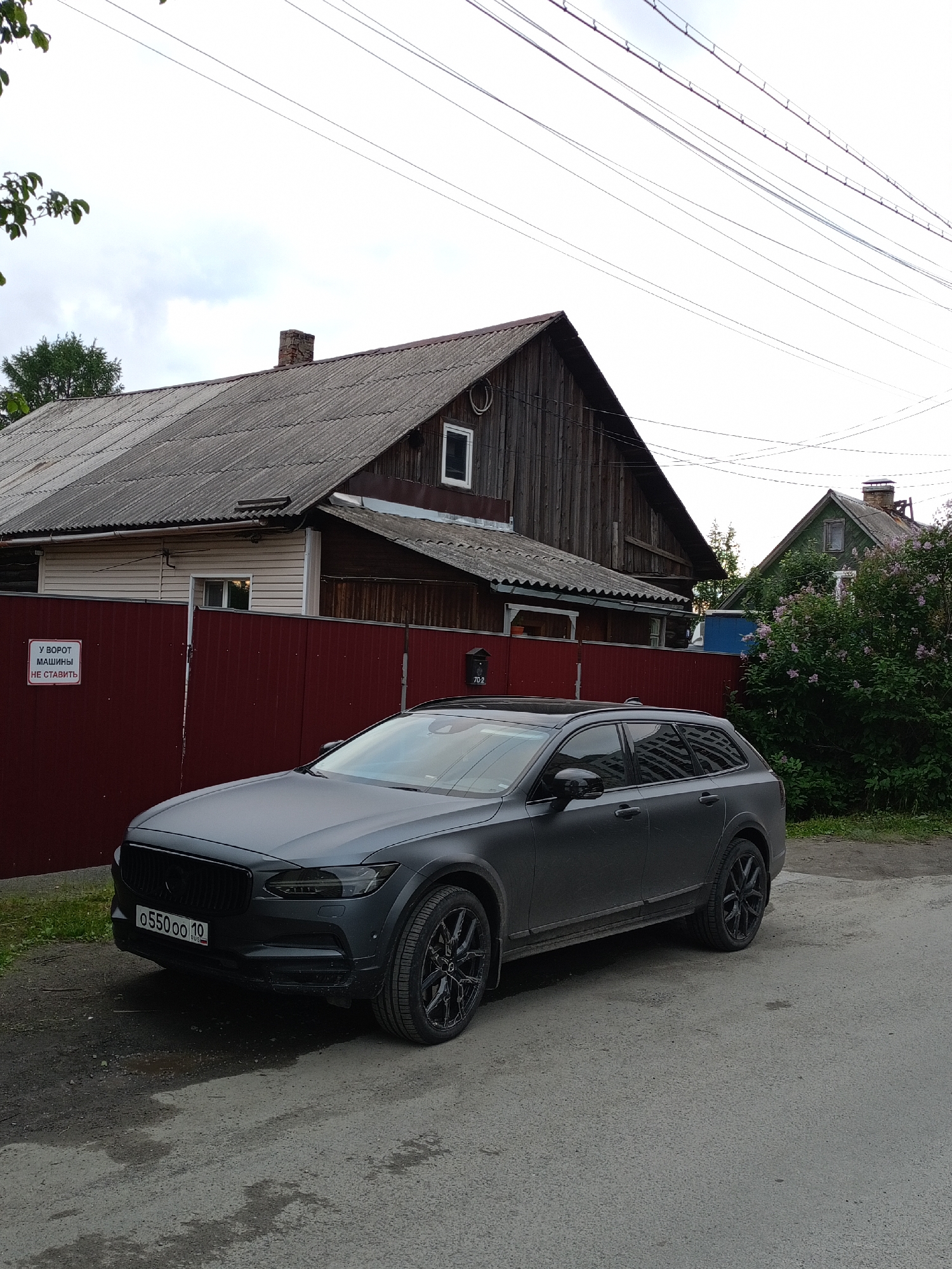 A dark gray Volvo station wagon is parked on an asphalt road in front of a weathered wooden house with a corrugated metal roof. A red fence runs along the property line, and a sign on the fence reads "У ВОРОТ МАШИНЫ НЕ СТАВИТЬ" (Do not park cars at the gate). A mailbox is attached to the fence. The sky is overcast, and trees and bushes are visible in the background. The license plate of the car is visible and reads "05500 10". The scene appears to be a residential area in Petrozavodsk, Russia.
