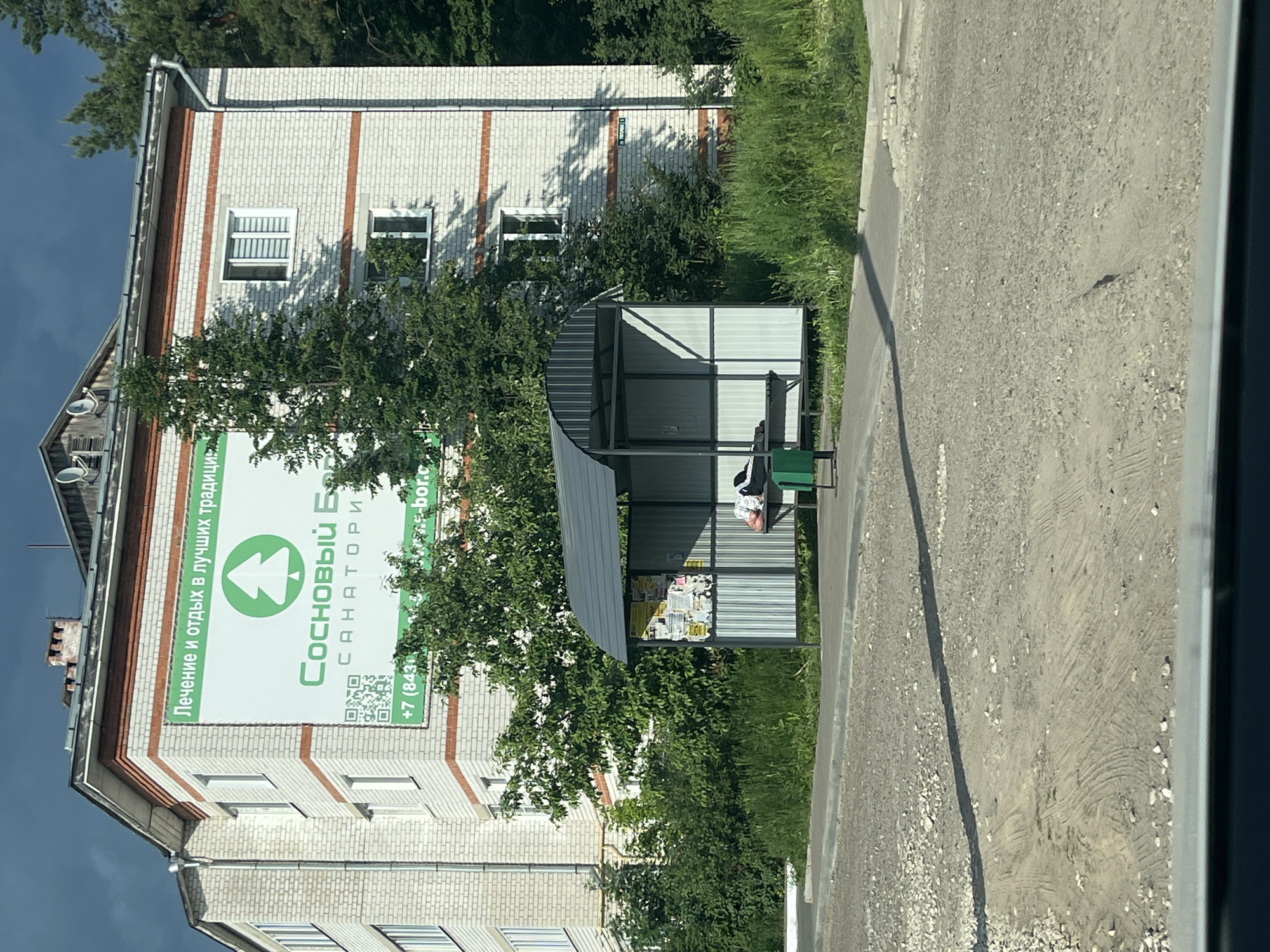 A bus stop shelter is in the foreground, with a person lying down on the bench inside. Behind the bus stop is a building with a large green advertisement on its facade. The advertisement features text in Russian and a green tree logo. To the right of the building, there are trees and foliage. The ground in front of the bus stop is a mix of asphalt and gravel. The scene appears to be in an outdoor urban or semi-urban environment. The advertisement on the building states "Лечение и отдых в лучших традициях" (Treatment and rest in the best traditions) and the name "СОСНОВЫЙ БОР" (Pine Forest), along with a phone number and website. It is likely located in Vasilievo, Russia, as indicated by the specific context of the advertisement.