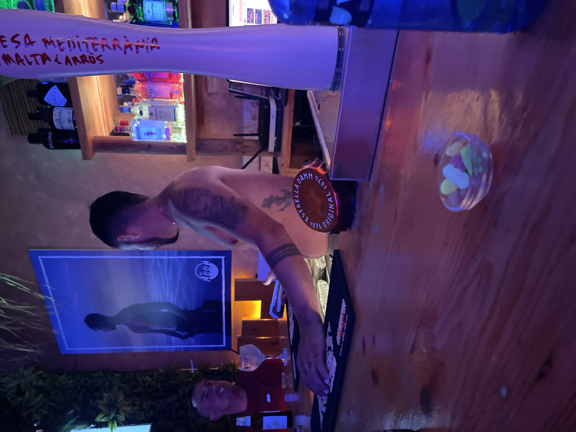 The image depicts an indoor scene at a bar, likely in Barcelona, Spain, given the prominent "ESTRELLA DAMM ORIGINAL 1876" label on a coaster, a well-known Spanish beer brand. A bartender, with tattoos on his arm and chest, is seen from the chest up, behind a wooden bar counter. He is wearing a black wristband. In the foreground, his hands are on the bar, possibly preparing or serving drinks. A small glass bowl with colorful gummy candies is also visible on the counter. To the left, a patron with short hair and a red shirt is partially visible, looking towards the bartender. Behind the bartender, shelves are stocked with various bottles of liquor and what appears to be a beer tap. A poster featuring a silhouetted male figure against a blue background is displayed on the wall behind the patron. The overall ambiance suggests a lively bar environment with dim, colored lighting.