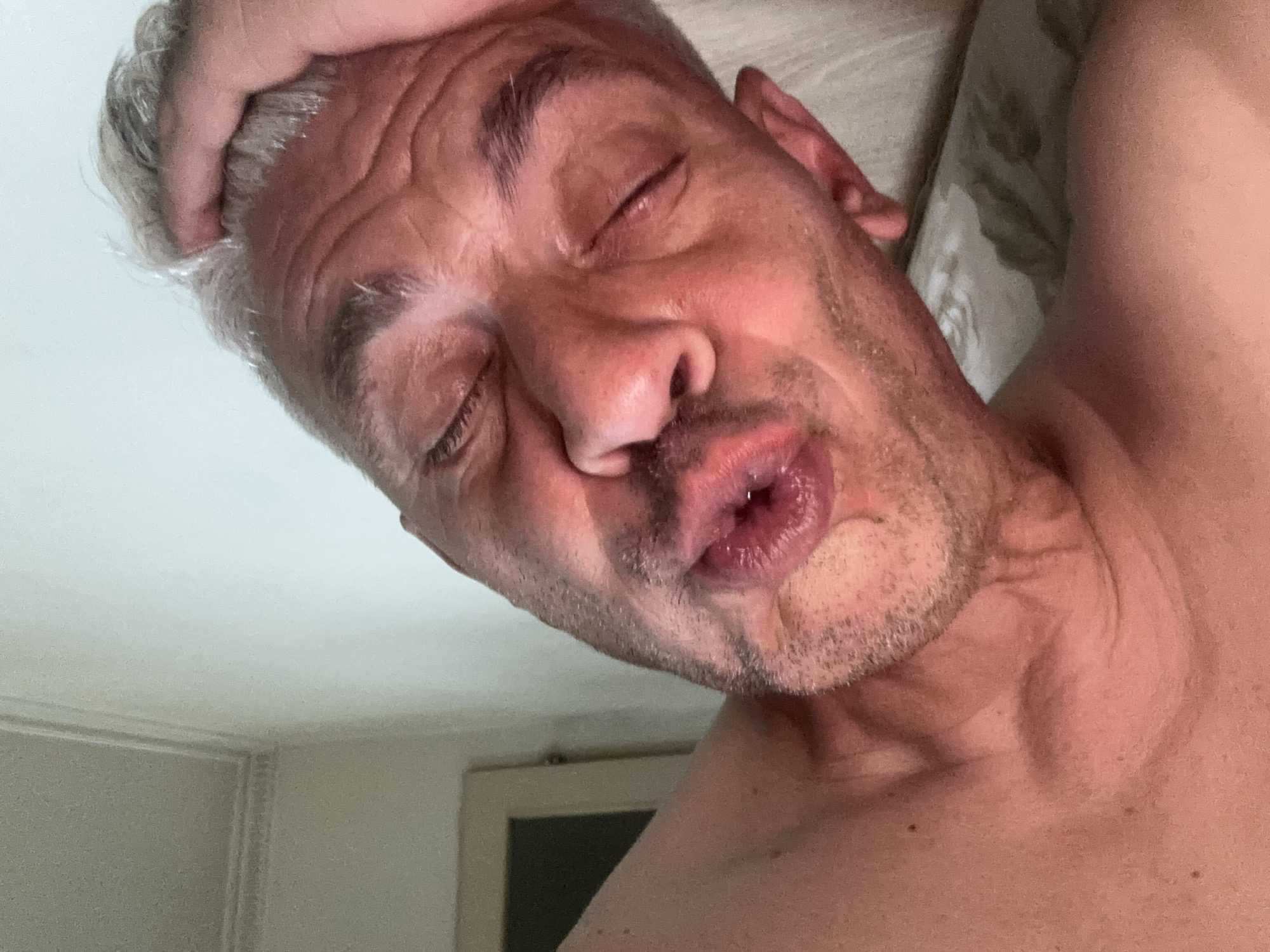 A close-up, angled portrait features a shirtless, middle-aged to older Caucasian male with grey hair, lying down in an indoor setting. His eyes are closed, and his lips are pursed in a slight pucker. Visible facial features include pronounced wrinkles on the forehead and around the closed eyes, along with light stubble around the chin and mouth. A hand, likely his own, with visible knuckles, is placed on the top of his head, partially obscured by his grey hair. The subject's left shoulder and upper chest are visible. In the background, to the upper left, a light-colored wall with white decorative molding is visible, suggesting architectural detail. A dark rectangular shape, possibly a door or mirror frame, can be faintly seen in the far left background. A textured, light-colored fabric, possibly bedding or a pillow, is present behind the subject's right side. The overall lighting appears soft and warm, casting subtle shadows.
Location: Unknown, Unknown.
