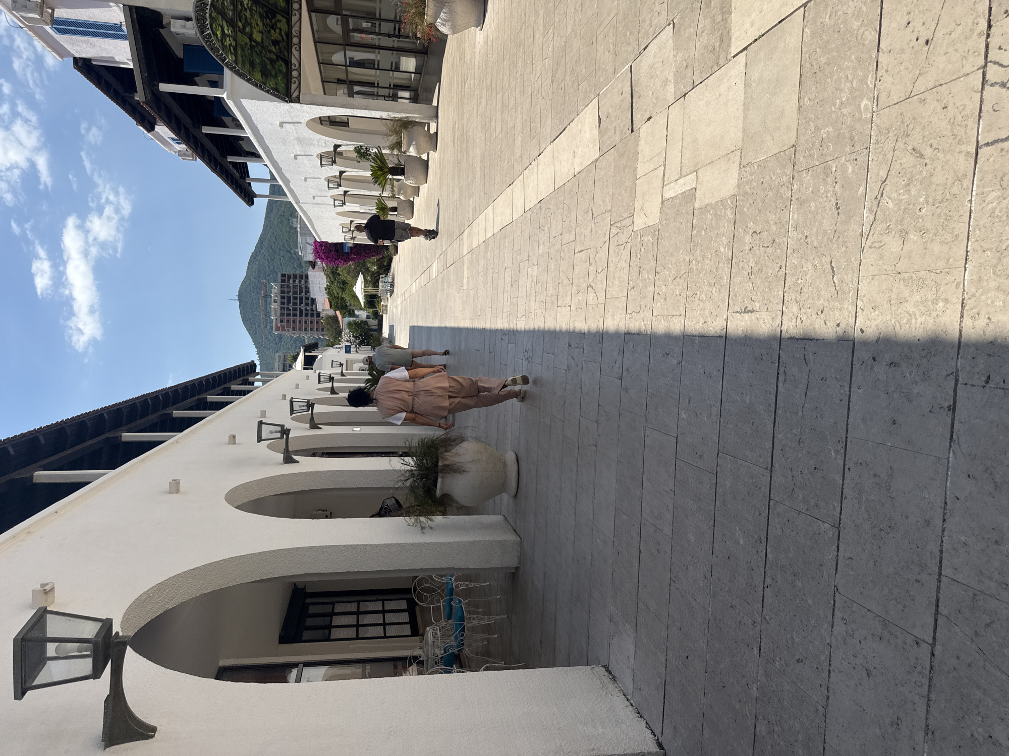A person in a light brown outfit is walking away from the camera down a sunlit paved walkway lined with white stucco buildings. The buildings feature arched entrances, and some have decorative outdoor lighting fixtures. Further down the walkway, another person in darker clothing is visible. In the background, a lush green mountainside rises under a bright blue sky with scattered white clouds. A partially constructed building is visible on the slope of the mountain, suggesting ongoing development in the area. The scene appears to be a pedestrian area in a Mediterranean-style town, likely a resort or commercial district, given the architecture and the presence of outdoor seating areas with white metal chairs and tables. The lighting suggests it is a sunny day, possibly late morning or early afternoon. The overall atmosphere is tranquil, with the focus on the architecture and the natural landscape. There is no visible text in the image.