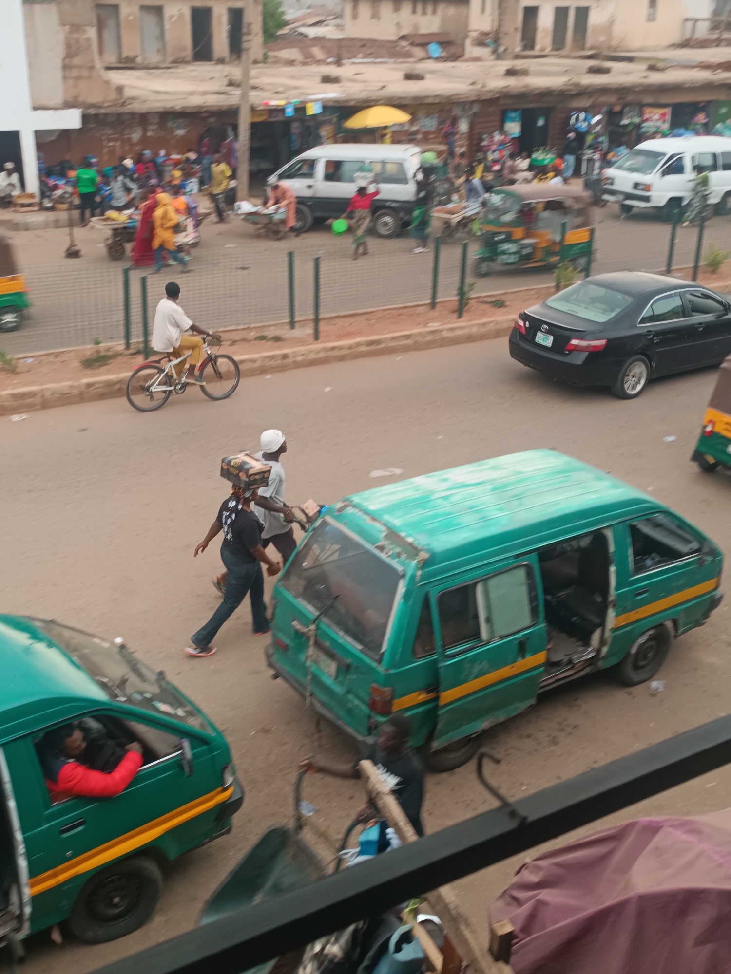 The image shows a street scene in Bukuru, Nigeria. Several people are walking and cycling on the road. There are a variety of vehicles including green minibuses, a black car, a white van, and auto rickshaws (tuk-tuks). One man is cycling on a bicycle. Two men are walking, one of whom is carrying a box on his head. One green minibus has its door open. There is a busy scene in the background with shops and people. The buildings in the background are old and appear to be commercial establishments. A fence separates the sidewalk from the road. There is an overcast sky.