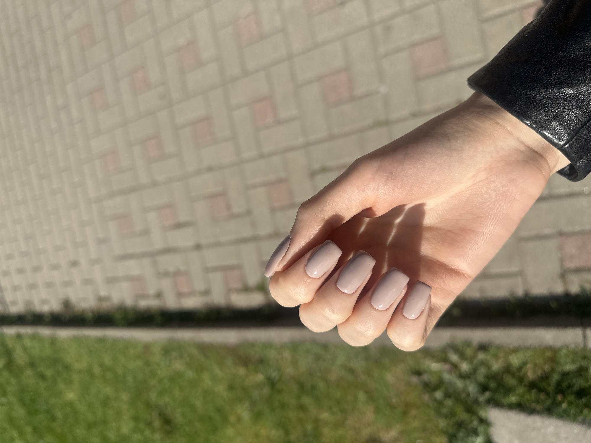 A close-up outdoor shot captures a hand adorned with light beige, squoval-shaped manicured nails, held slightly cupped against a sunlit background. The hand appears to be resting or posed to showcase the manicure. The cuff of a black leather jacket is visible around the wrist, indicating the person is wearing outerwear.
The background is sharply divided into two main sections: an upper area of light-colored paving stones and a lower strip of vibrant green grass. The paving features a distinctive pattern of rectangular greyish-beige blocks interspersed with smaller, reddish-pink square tiles, suggesting a sidewalk or public plaza. The grass strip, slightly out of focus, separates the paved area from an unseen further distance.
Bright, direct sunlight illuminates the scene, casting prominent shadows from the fingers onto the palm of the hand and creating strong contrasts. This lighting suggests a clear day, possibly morning or late afternoon, given the angle of the shadows. There is no discernible text in the image. The setting is an urban or suburban outdoor environment, consistent with the provided location context of Sofia, Bulgaria.