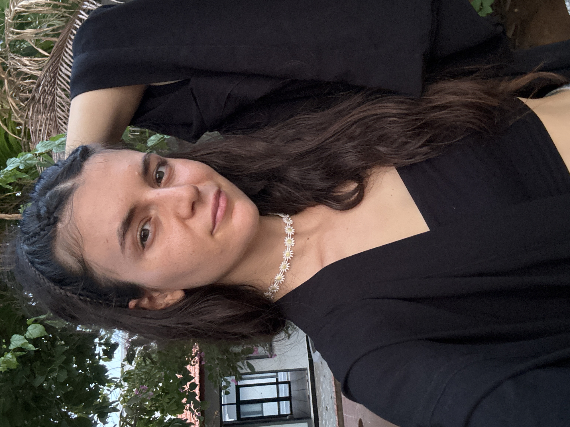 A young woman is depicted in a selfie-style portrait, looking directly at the camera with a subtle, gentle smile. She has long, dark brown, wavy hair, parted in the middle, with small braids framing her face. Her face features freckles, and she has a relaxed, pleasant expression. She is wearing a black, long-sleeved top or dress with a square neckline, and a white and yellow daisy-chain choker necklace adorns her neck.
The photograph is taken outdoors, set against a backdrop of vibrant green foliage, indicative of a tropical environment. Large palm fronds are visible in the upper left of the frame, and various other leafy plants, some with small purple flowers, fill the lower left. In the mid-ground, behind the woman's right shoulder, there are architectural elements, possibly part of a building or balcony, with dark-framed windows reflecting the bright sky.
The lighting is natural and soft, suggesting it is daytime, possibly late morning or early afternoon, given the diffused illumination and lack of harsh shadows. The overall mood is calm and serene. The tropical vegetation is consistent with the provided location context of Mũi Né, Vietnam. No visible text is present in the image.