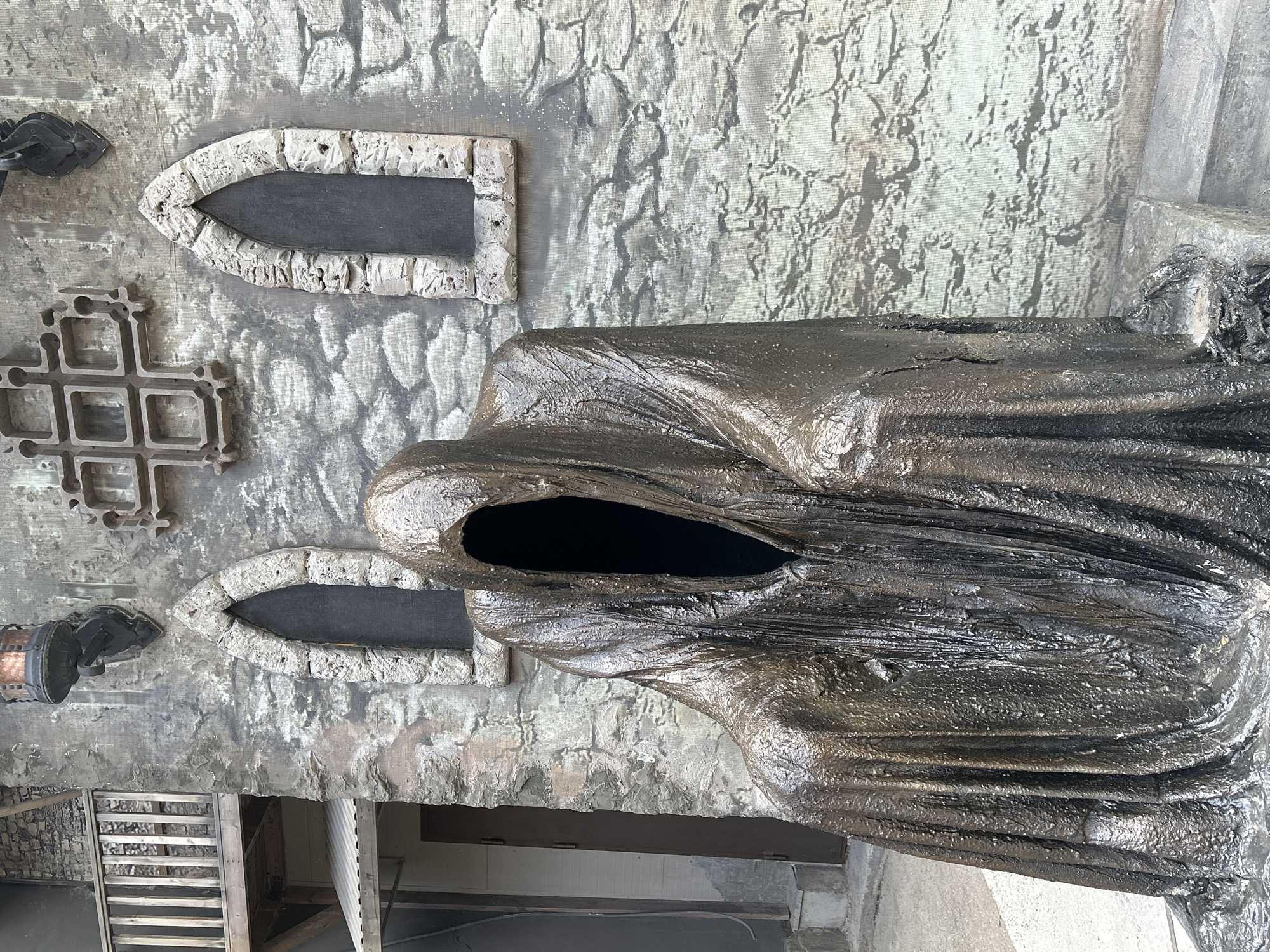 An outdoor, full-shot, eye-level, medium-wide photograph shows a sculpture of a cloaked, shadowy figure with a vacant opening for a face, sitting in front of a faux stone wall designed to resemble a medieval building. The sculpture is dark brown and appears to be made of metal, with a heavily textured, dripping surface. The wall behind it is grey and textured to look like rough-cut stones. There are two pointed, arched windows on the wall, filled with dark material. Above the windows, a decorative wrought-iron grille is mounted. A rustic wooden balcony with railings is visible on the left side of the image, attached to a lighter-colored building. A dark brown door is also visible on the left. The scene is lit by natural daylight, casting subtle shadows. The location is identified as the city of Perm, Russia.