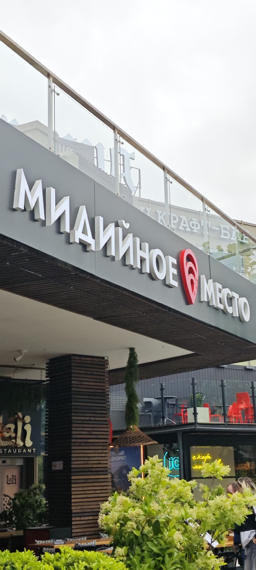 The image depicts the exterior of a restaurant named "Midyaynoye Mesto," which translates to "Mussel Place," located in Sochi, Russia. The restaurant's signage is prominently displayed with white Cyrillic letters and a red location pin graphic. The building features a modern architectural design with a glass railing on an upper level.
In the foreground, there are trees with white flowers. Other nearby businesses are visible, including "Leli Restaurant" and another with neon signage in Russian. Tables and chairs are visible outside the restaurants, suggesting an outdoor dining area. The overall environment appears to be an urban commercial district, likely a tourist area.