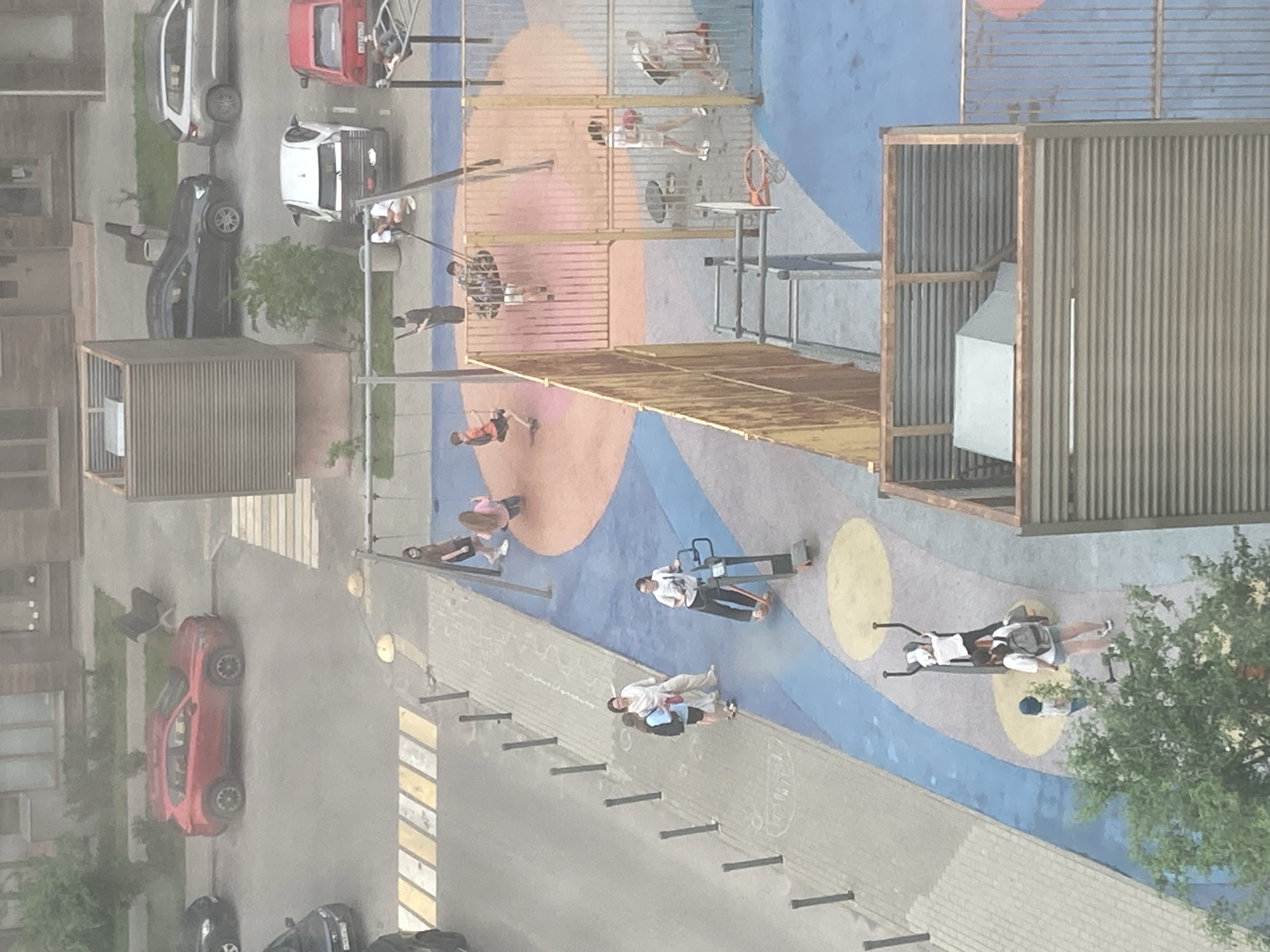 Captured from an elevated perspective, the image shows a playground and adjacent street scenes in Khimki, Russia. The playground, rendered in various colors, features equipment occupied by children and adults engaged in play and physical activity. Some individuals are on swings and climbing structures. The surrounding street has parked vehicles, including cars of red and white hues. Pedestrians can be observed on the walkway. The environment includes urban infrastructure such as buildings and vegetation, suggesting a residential area. The lighting indicates daytime conditions, although the image has a slightly hazy or muted quality.