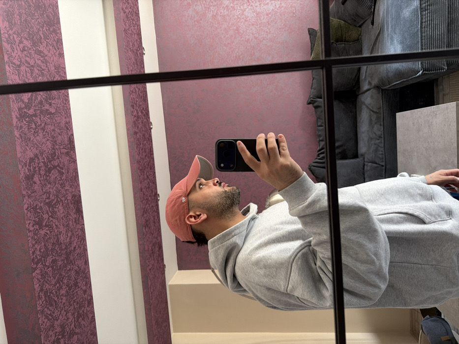 A male individual is depicted in a mirror selfie taken within an indoor residence in Sofia, Bulgaria. The subject, visible from the mid-torso up, wears an oversized light grey hoodie with a front pouch and a faded pinkish-orange baseball cap featuring a '47' logo and a light grey underside brim. He has a beard and mustache. His right hand holds a dark-colored smartphone, consistent with an iPhone's multi-lens design, angled towards the mirror to capture his reflection. The mirror itself has a dark, grid-patterned frame. The reflected background reveals walls with two distinct horizontal bands of textured dark pink/burgundy wallpaper, separated by a plain white strip. A white ceiling with recessed lighting is visible. To the right, a dark grey sectional sofa with ridged upholstery is present, adorned with several dark grey pillows and one olive green throw pillow. In front of the sofa, a rectangular light grey coffee table stands. A light-colored bag is partially visible on the floor to the far left.