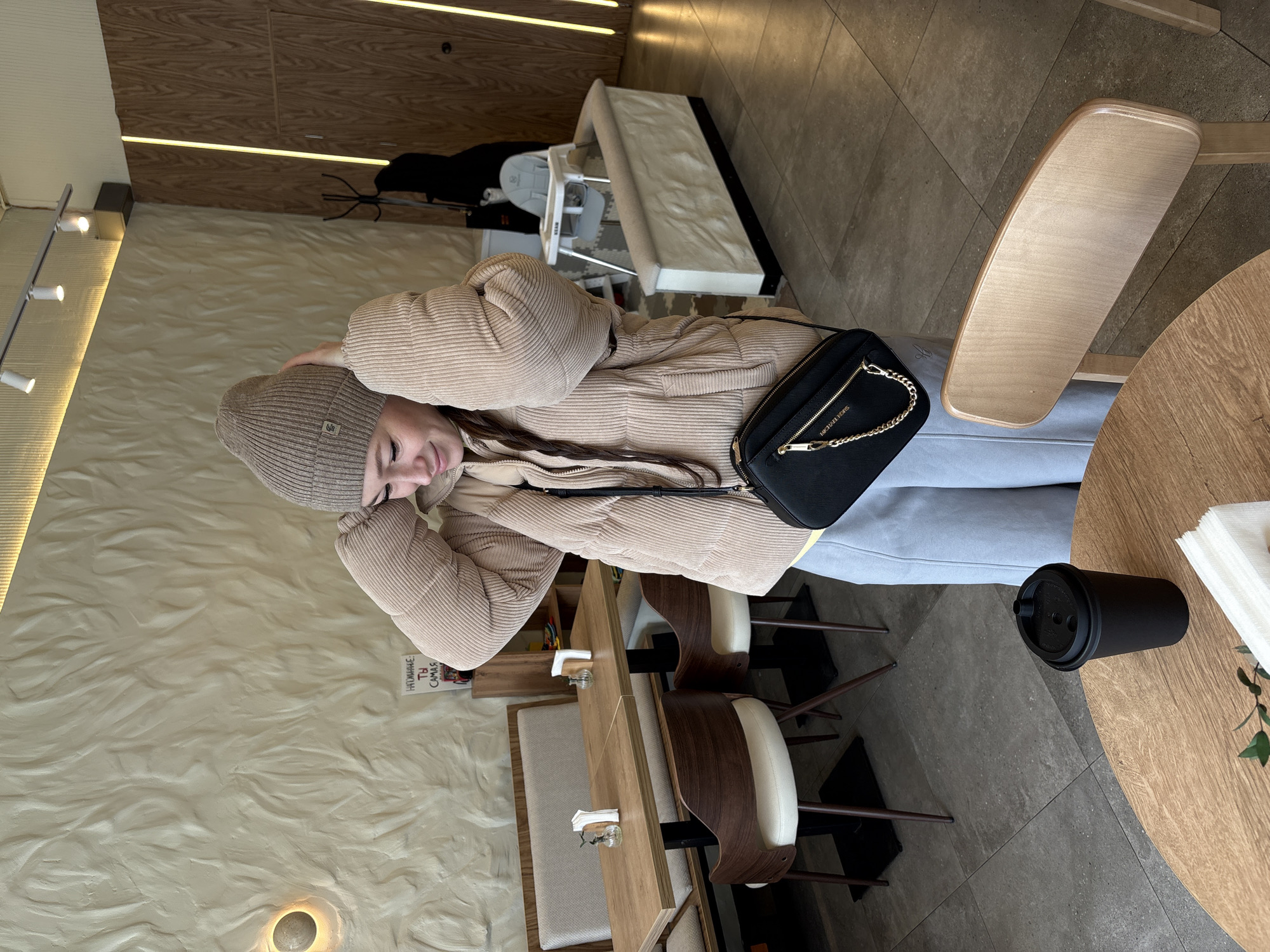 A young woman wearing a beige beanie and a beige corduroy jacket is sitting at a wooden table in an indoor cafe. She has a black purse with a gold chain strap hanging over her shoulder. A black to-go coffee cup is on the table beside her. The cafe has a light-colored wavy patterned wall and wooden paneling with built-in lighting. There are wooden chairs and a wooden bench with beige cushions. A high chair and a crib are visible in the background. The cafe appears to be located in Ulyanovsk, Russia, based on the text in Cyrillic on an object on the table next to the woman's head.