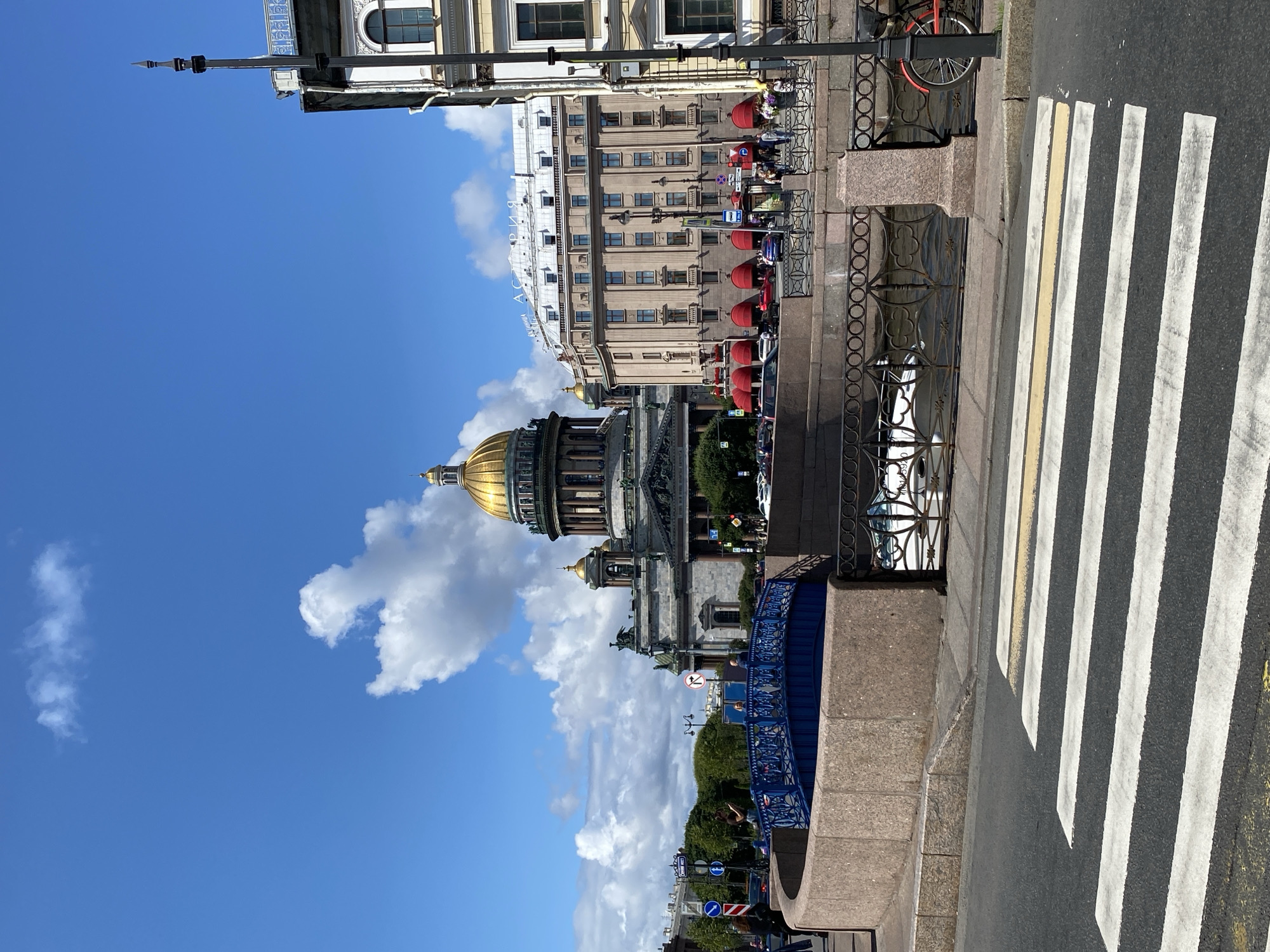 This is a daytime, eye-level, exterior shot of St. Isaac's Cathedral in Saint Petersburg, Russia, with a focus on a canal, a pedestrian bridge, and the surrounding architecture. In the foreground, a zebra crossing is visible on an asphalt road. Behind the zebra crossing, a concrete embankment lines a canal, with a decorative metal railing above it. A bright blue pedestrian bridge with intricate metalwork spans the canal.
In the middle ground, St. Isaac's Cathedral, with its prominent golden dome, is a central architectural feature. Adjacent to the cathedral is a large, light-brown building with many windows and red awnings over some of its ground-floor entrances. The building has a sign on its roof that reads "АСТОРИЯ" (Astoria).
Several people are present: some are walking across the bridge, others are on the sidewalks near the buildings, and a few individuals appear to be gathered near the red awnings. Cars are parked and driving on the street. A red bicycle is parked next to a lamppost on the right side of the image.
The environment is urban, with trees visible in the background. The sky is blue with scattered white clouds. The lighting suggests a sunny day.