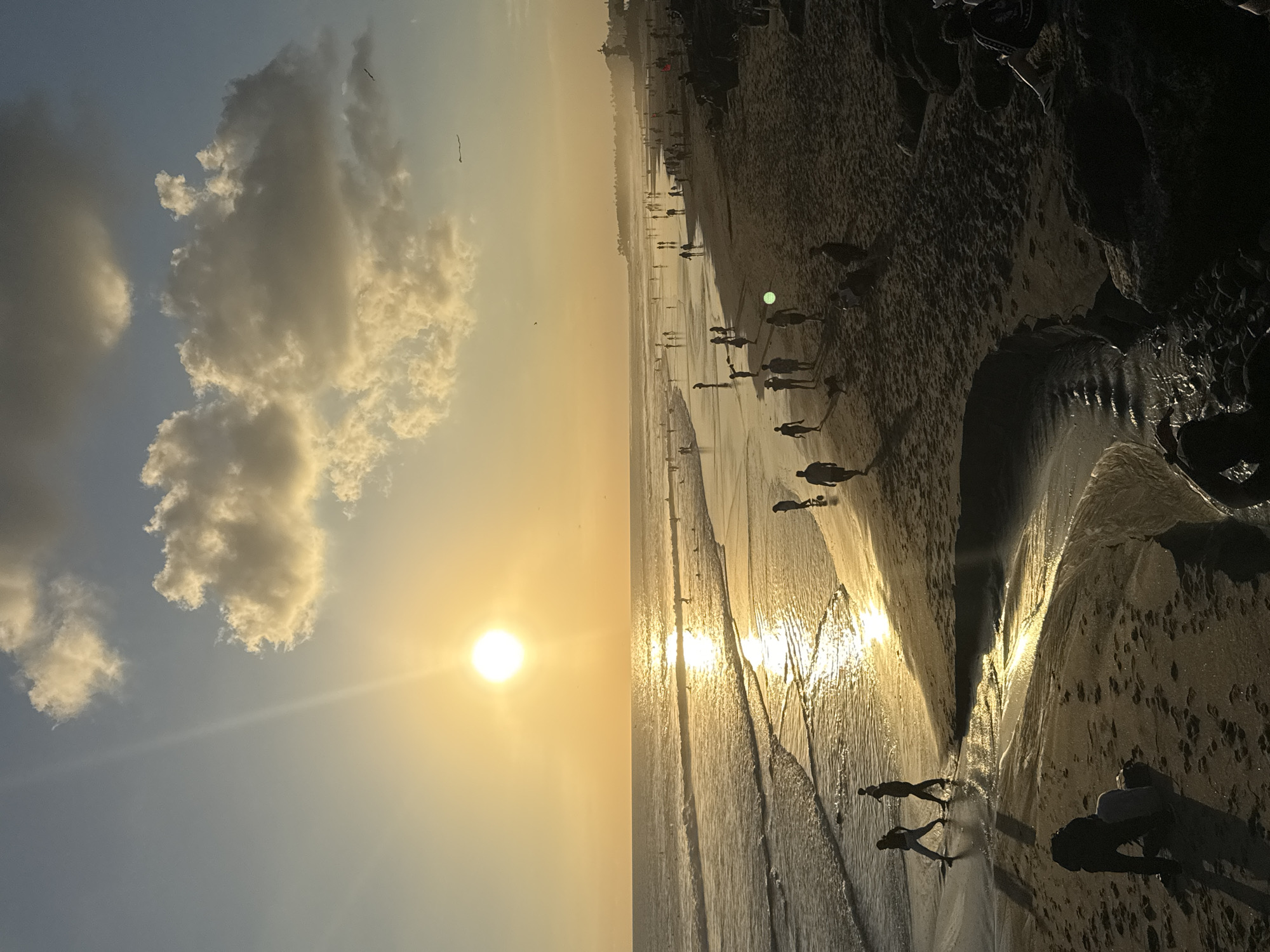 The image shows a beach scene at sunset in North Kuta, Indonesia. The sun is positioned above the horizon, casting a golden glow over the ocean and beach. The sky is a gradient of blue to yellow near the horizon, with puffy white clouds in the upper left. People are walking along the shoreline, their silhouettes visible against the setting sun. The beach itself transitions from sand to rocky formations towards the right side of the image. The water reflects the sunlight, creating shimmering patterns.