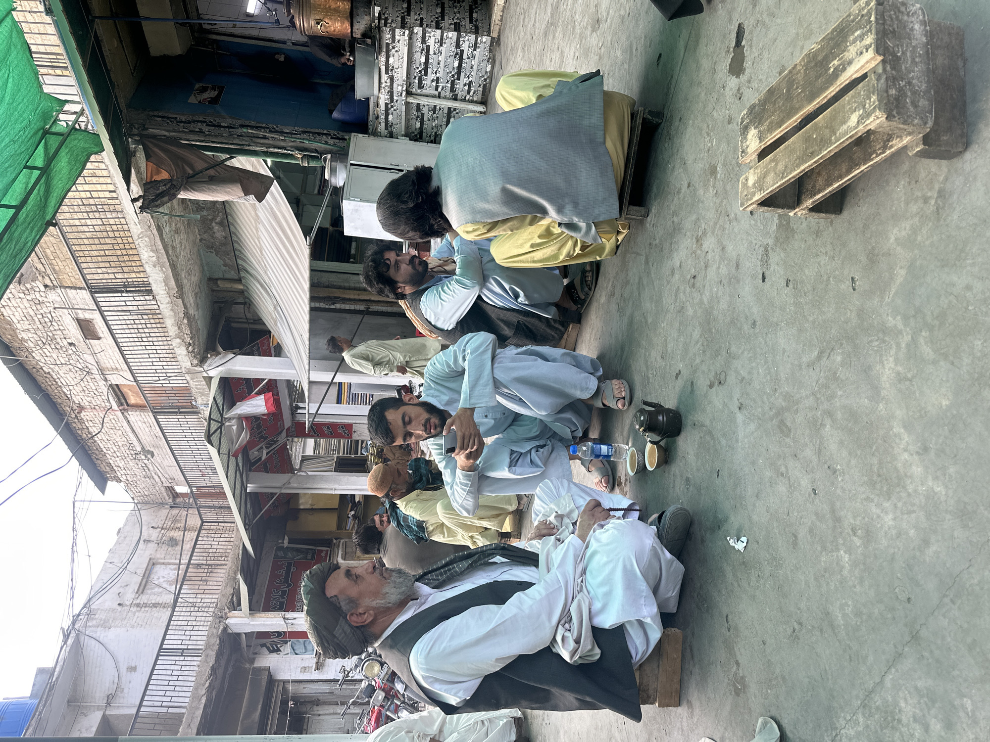 In Quetta City Tehsil, Pakistan, a group of men sit in a line on low wooden stools or pallets on a paved surface. The men appear to be conversing and drinking from small cups and a teapot is visible. They are dressed in traditional Pakistani clothing, including shalwar kameez and vests. The setting is an urban area, with buildings and awnings visible in the background.