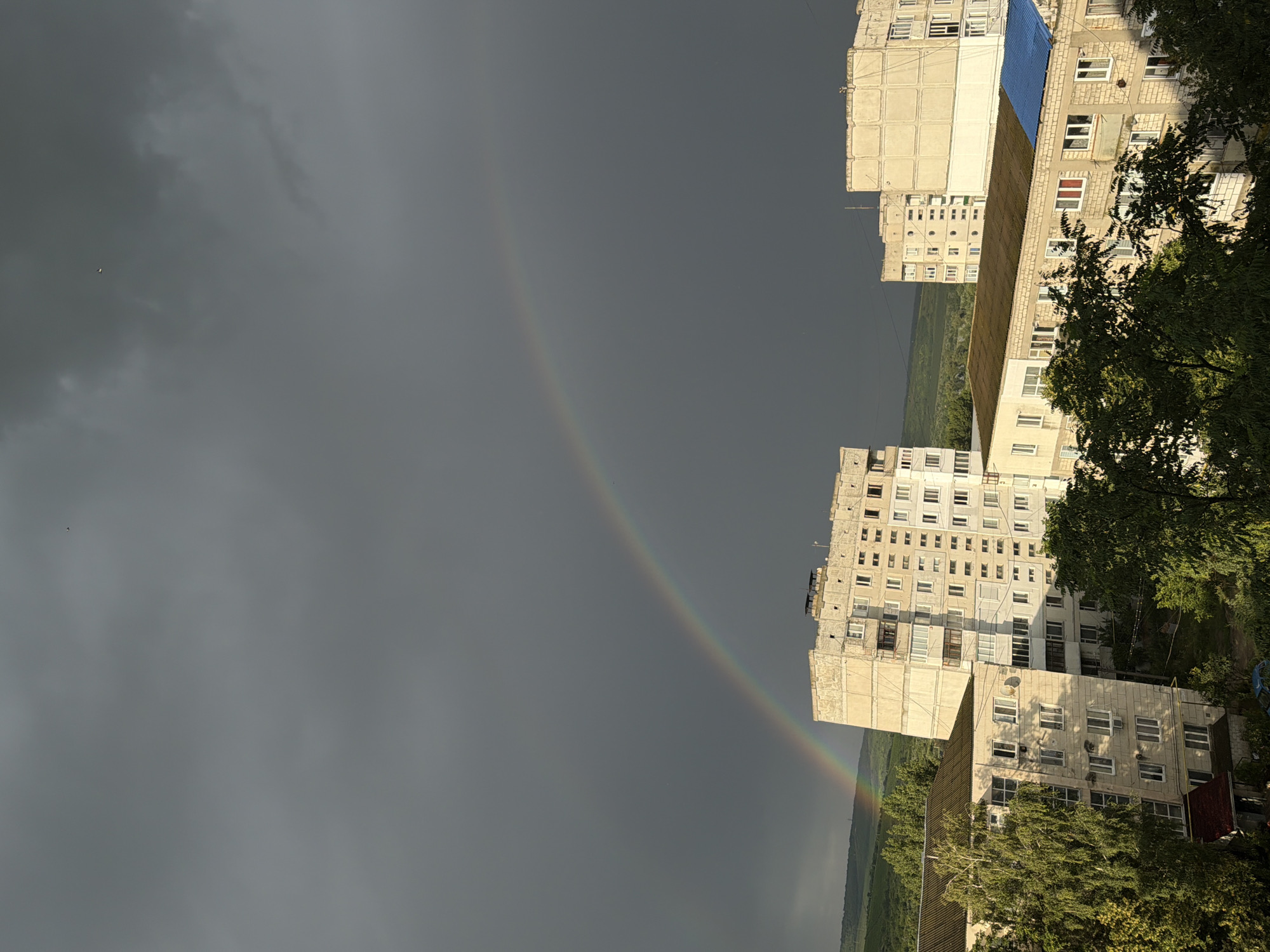 An aerial view captures a dramatic sky over the city of Strășeni, Moldova, with a prominent rainbow arcing across the dark, stormy clouds. The cityscape below features several multi-story apartment buildings, characteristic of Soviet-era architecture, with visible windows and balconies. Lush green trees and rolling hills form the background, suggesting a semi-rural or suburban setting within the city. The lighting indicates either early morning or late afternoon, with the sun breaking through the clouds to cast a spotlight on parts of the buildings. A single small dark speck, possibly a bird or a distant aircraft, is visible in the upper right quadrant of the sky.