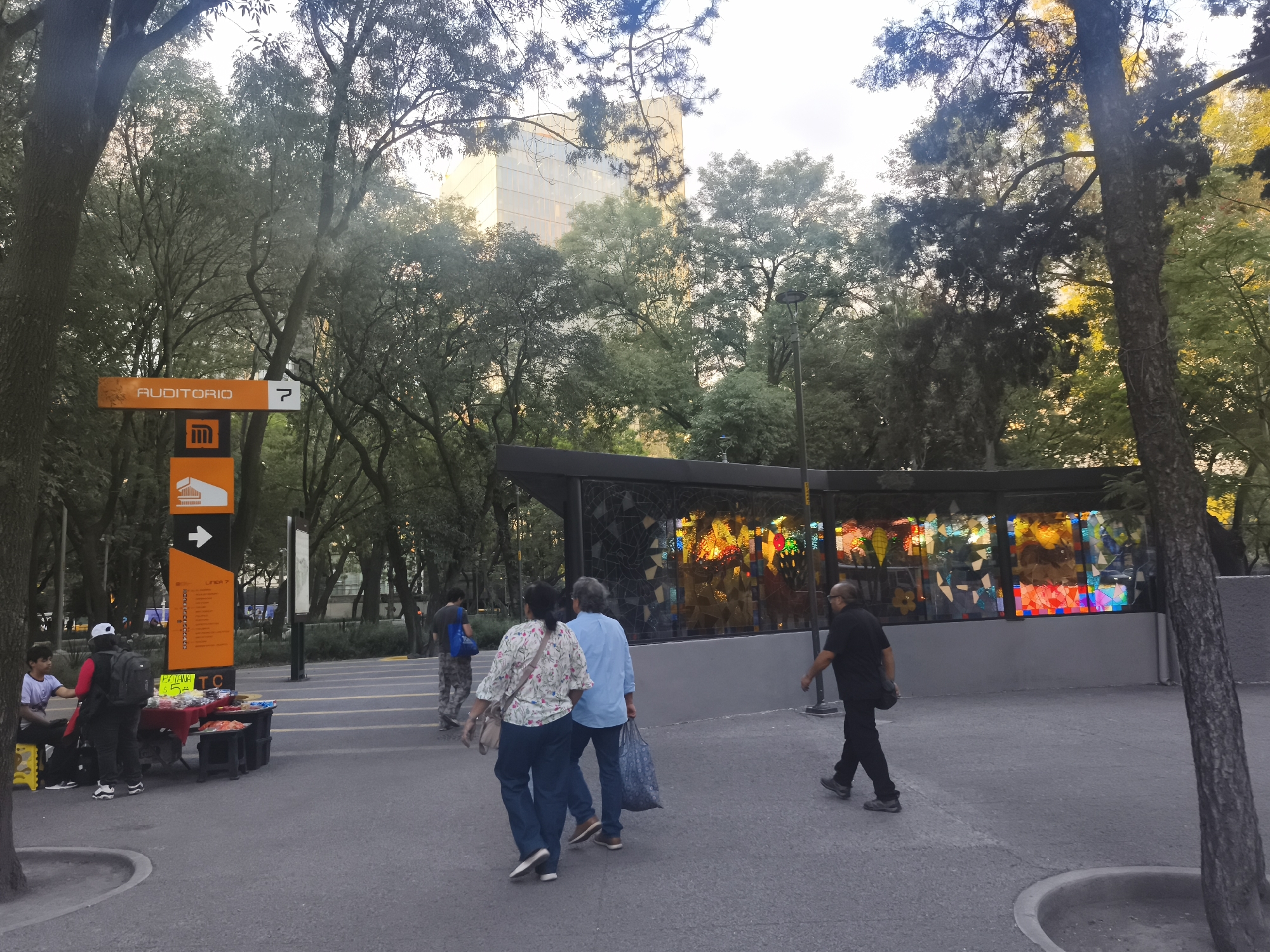 This image depicts a bustling outdoor scene in Mexico City, specifically near the Auditorio Nacional Metro station. The time of day appears to be late afternoon or early evening, with a soft, warm light filtering through numerous green trees that fill the background and frame the scene.
In the foreground, a paved path is occupied by several individuals. On the left, a street vendor operates a small stall. Visible items include what appear to be snacks, with a sign partially reading "PLATANOS" and a price. A person is seated next to the stall, and another individual, possibly a customer or helper, stands nearby.
A prominent orange and black sign stands tall on the left, clearly marking "AUDITORIO" and the number "7", accompanied by the iconic Mexico City Metro logo. A smaller panel below it also indicates "LINEA 7" with a directional arrow.
In the midground, several pedestrians are walking. A group of three individuals walks away from the camera towards the right side of the path. Behind them, a striking structure with colorful, stained-glass-like windows creates a vibrant focal point, characteristic of art installations often found at metro stations. Another person, dressed in black, walks past this colorful structure from right to left.
Further in the background, a modern building with a light-colored facade can be seen peeking above the tree line, confirming the urban environment. The scene captures a typical moment of daily life around a public transit hub, with people commuting and street vendors operating.
