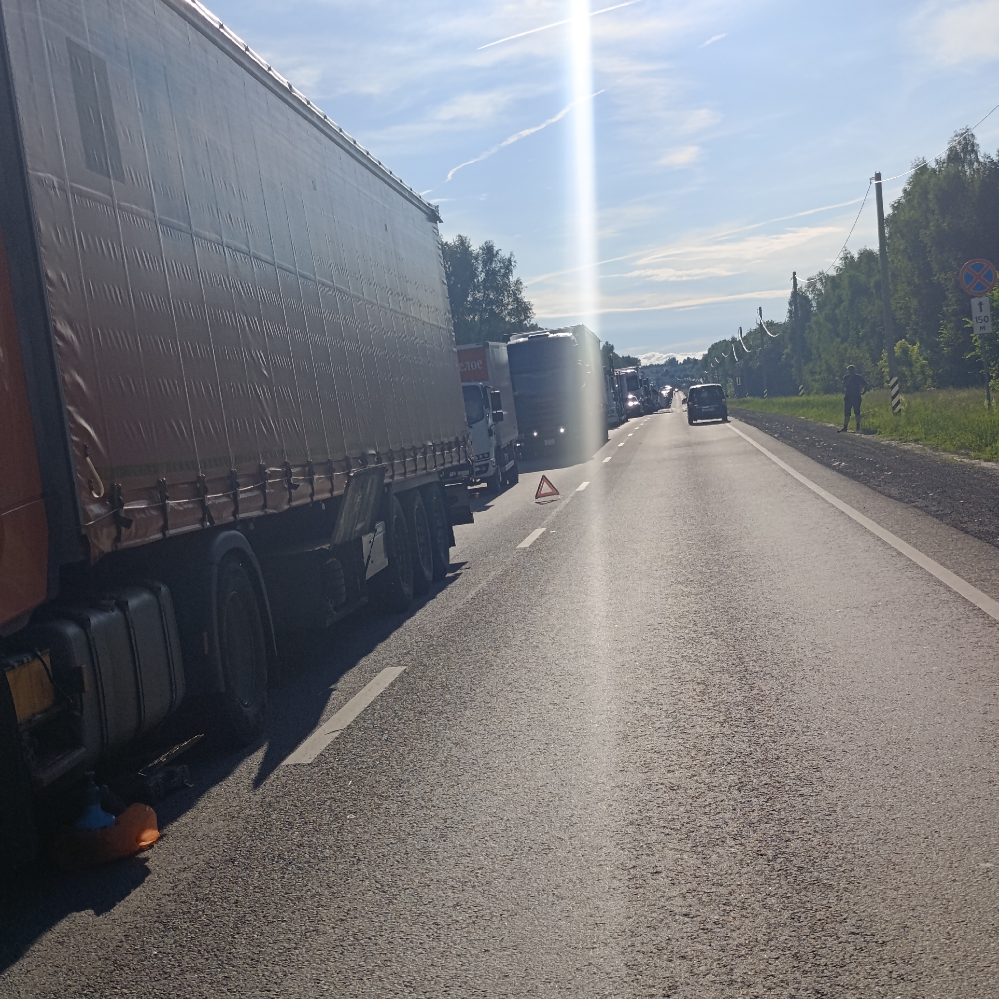 In L'vovo, Russia, on a sunny day with a clear blue sky and scattered contrails, a traffic jam is depicted on a two-lane asphalt road. The foreground is dominated by the side of a large, beige-covered semi-trailer truck, its tarp exhibiting vertical pleats and secured with horizontal straps. The truck has multiple axles and wheels visible. To the right of the truck, the road is lined with other vehicles, including several semi-trailer trucks and a dark-colored passenger van in the distance. An orange traffic cone is visible near the front wheel of the foreground truck, and a red triangular warning sign is placed on the road between the truck and the oncoming traffic. Further down the road, a lone figure stands on the right shoulder, facing away from the camera. The shoulder is covered in gravel and bordered by a dense line of green trees. A road sign with a red circle and a blue symbol is posted on the right side of the road, with text indicating "150 M". Overhead power lines with insulators are strung along the side of the road, supported by wooden poles. The scene suggests a stalled or slow-moving traffic situation on a rural or suburban highway.