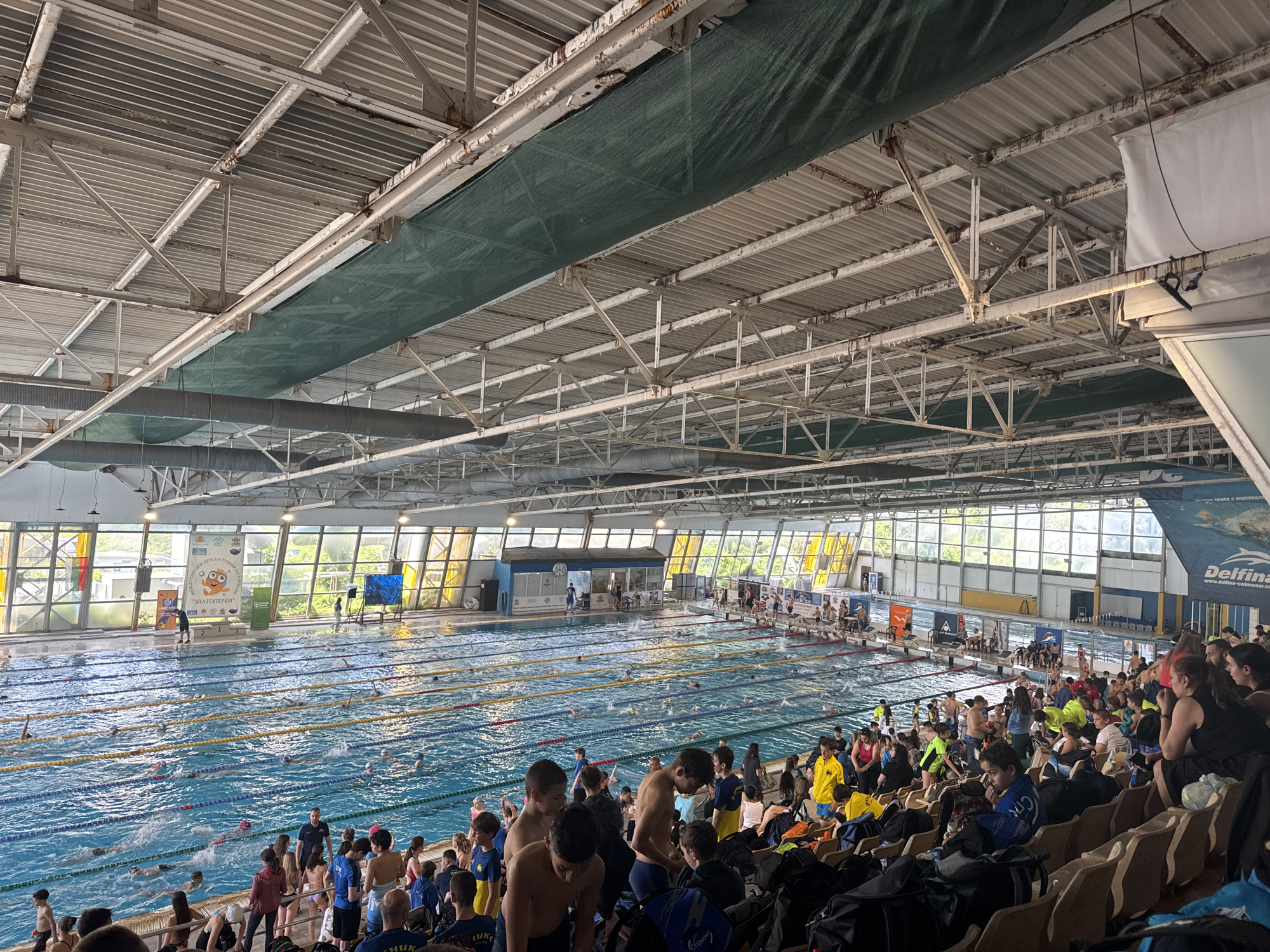The image shows an indoor swimming pool facility. Several swimmers are in the pool, divided by lane ropes. Spectators are seated in stadium-style seating to the right. Many of the spectators are young and appear to be swimmers or team supporters. Other people are standing around the edge of the pool. The facility has a high ceiling with exposed structural elements and ventilation ducts. Daylight enters through large windows along the side of the pool. Banners and signage are visible in the background. This swimming pool facility appears to be the "Delfina" swimming complex in the city of Sofia, Bulgaria.