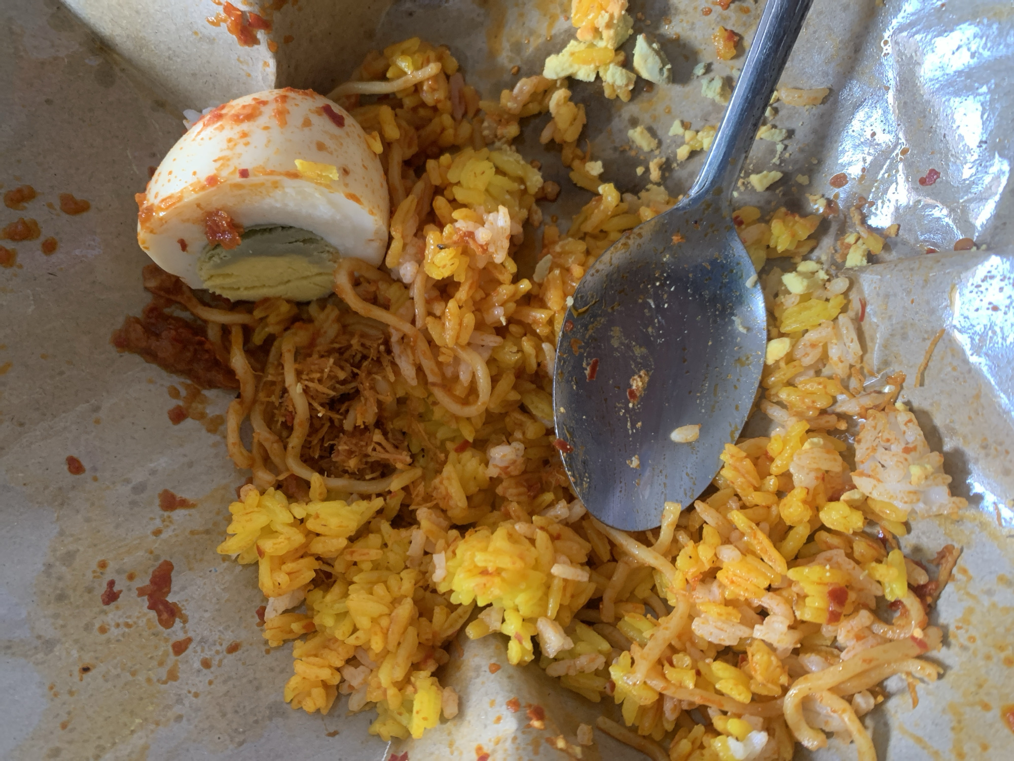 The image shows a close-up, overhead view of a portion of yellow rice with bits of egg and shredded meat, served on a piece of brown paper. A metal spoon rests on the rice, partially obscuring the food. The rice appears to be seasoned with spices, giving it a vibrant yellow and orange hue. There are specks of red chili flakes scattered throughout the dish. A hard-boiled egg, cut in half lengthwise, is positioned on the left side of the frame, revealing a pale yellow yolk and a white albumen. The egg is also sprinkled with red chili flakes. The paper packaging is creased and stained with traces of the food, suggesting it is a disposable container. The scene is likely from a food stall or a casual dining establishment, possibly in Sungai Meriam, Indonesia, where such street food is common. The lighting appears to be natural, coming from above, casting subtle shadows. There are no people visible in the image.