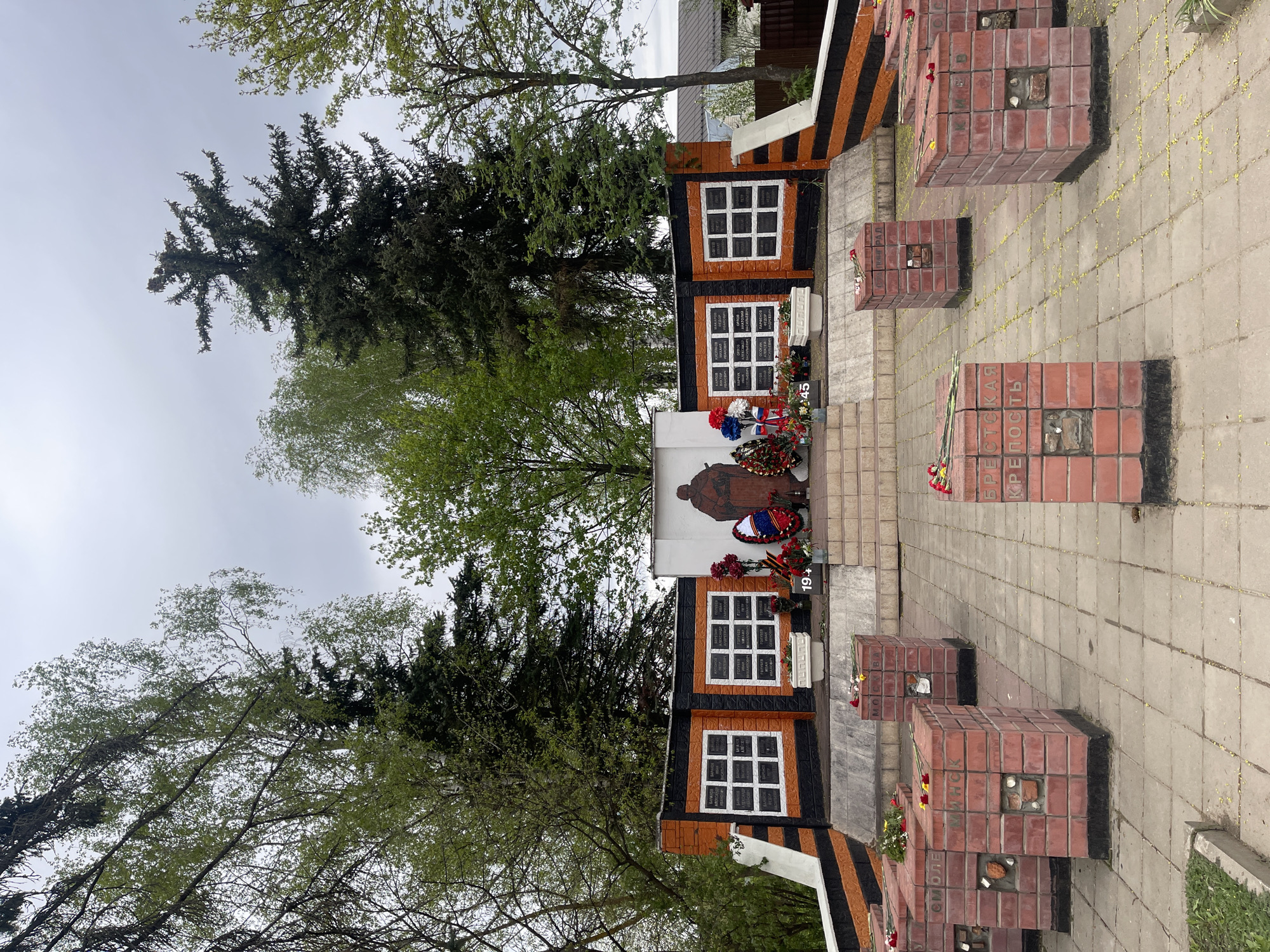 A monument is in an outdoor space in Хлябово, Russia. It consists of a white wall with a drawing of a soldier in an overcoat and a monument with two sets of six white rectangles on a black background. There are red brick structures with a single brick missing on top that are lined up in rows on the grey concrete. Wreaths and flower arrangements are placed on the monument and on the brick structures. Tall trees are around the monuments and buildings are on the background. The sky is grey.