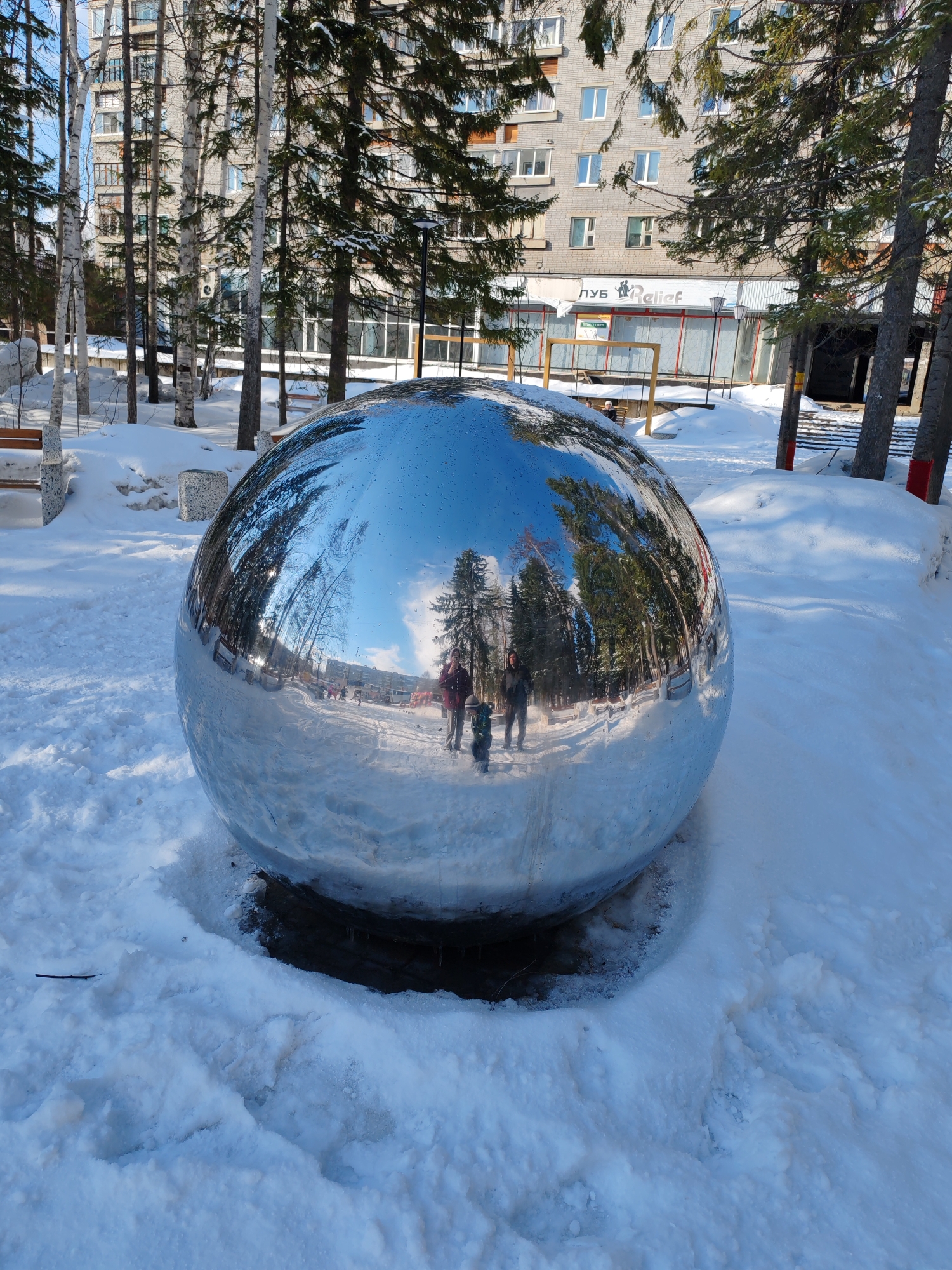 The image presents a winter scene in Ust-Ilimsk, Russia, featuring a large, reflective metal sphere resting in a snowy park. The sphere mirrors the surroundings, including evergreen and birch trees, a multi-story residential building, and a building with the Cyrillic inscription "ЛУБ Relief". In the sphere's reflection, two adults and a child are walking on the snow-covered ground. The ground is covered in snow, and the sphere sits in a small depression. Sunlight illuminates the scene, creating bright reflections on the sphere and casting shadows on the snow. A park bench is visible to the left.