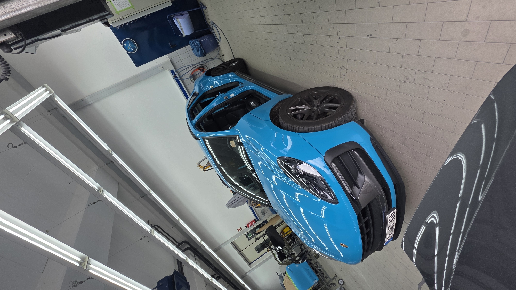 In a brightly lit indoor workshop, a vibrant cyan Porsche Macan with its doors open is parked on a light-colored tiled floor. The car is the central focus, with its sleek design and glossy finish reflecting the overhead fluorescent lights. Various automotive tools and equipment are visible in the background, including a blue cabinet with a clock, a roll of paper, and a blue trash bin. To the left of the car, another vehicle, possibly a car door, is covered with a light brown protective sheet and is on a wheeled stand. In the foreground, the hood of a dark gray car is partially visible, reflecting the overhead lighting in a striking pattern. The overall impression is that of a professional car detailing or repair shop.