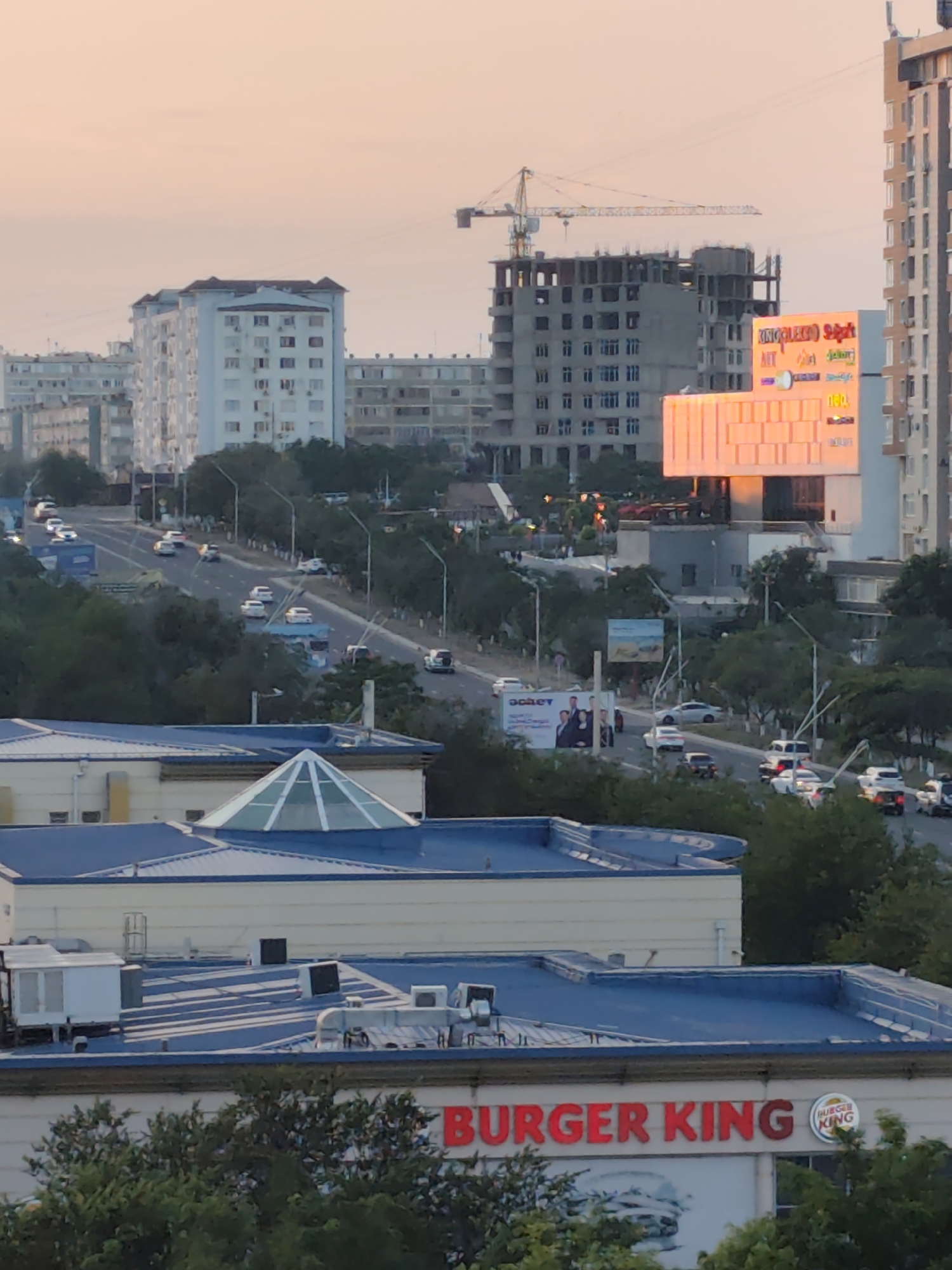 The image depicts a street scene in Aktau, Kazakhstan, likely during sunset, given the warm, diffused light and soft hues in the sky. In the foreground, the roofs of modern buildings are visible, one with a distinctive blue metal roof and a glass-domed skylight. Another building below features a prominent red sign reading "BURGER KING" with its logo. A large advertisement featuring a burger is also visible beneath the Burger King sign.
Further back, a multi-lane road with moderate traffic is present, with cars and vans driving in both directions. Trees line the sides of the road and form green patches throughout the scene. Several multi-story residential buildings, typical of Soviet-era architecture, are visible in the mid-ground, painted in light colors.
In the background, a construction site with a crane indicates ongoing development. Adjacent to it, a tall building displays a vibrant, illuminated advertisement with various text and logos, featuring warm orange and yellow tones. The overall impression is of an urban environment with a mix of residential, commercial, and construction areas, set against a backdrop of a soft, atmospheric sky. No individuals are clearly identifiable in the image.