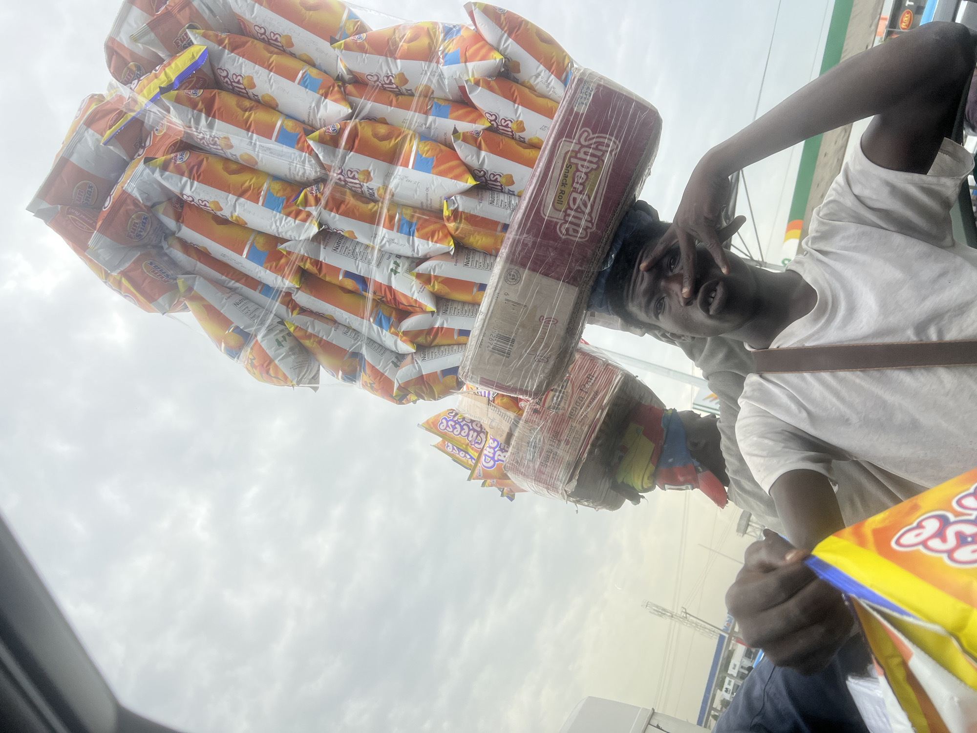 A street vendor in Igboefon, Nigeria, carries a large stack of "Smax" cheese-flavored snack bags and a "Super Bite Snack Roll" box on his head, wrapped in clear plastic. He is standing near a building with a "PLOT" sign, wearing a white t-shirt and a strap across his shoulder. He is holding another bag of "Smax" chips. The sky is overcast.