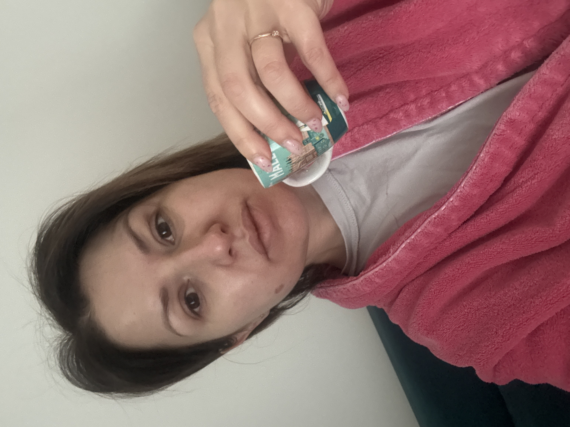 A woman is holding a small paper cup with a clear liquid in it. She is wearing a pink robe over a gray shirt. She has brown hair and light skin. Her nails are painted light pink with small pink polka dots. She is wearing a ring on her ring finger. The cup appears to be from "Mall of Sofia", suggesting the picture was taken in or near the Mall of Sofia shopping center in Sofia, Bulgaria. The background is a plain white wall.