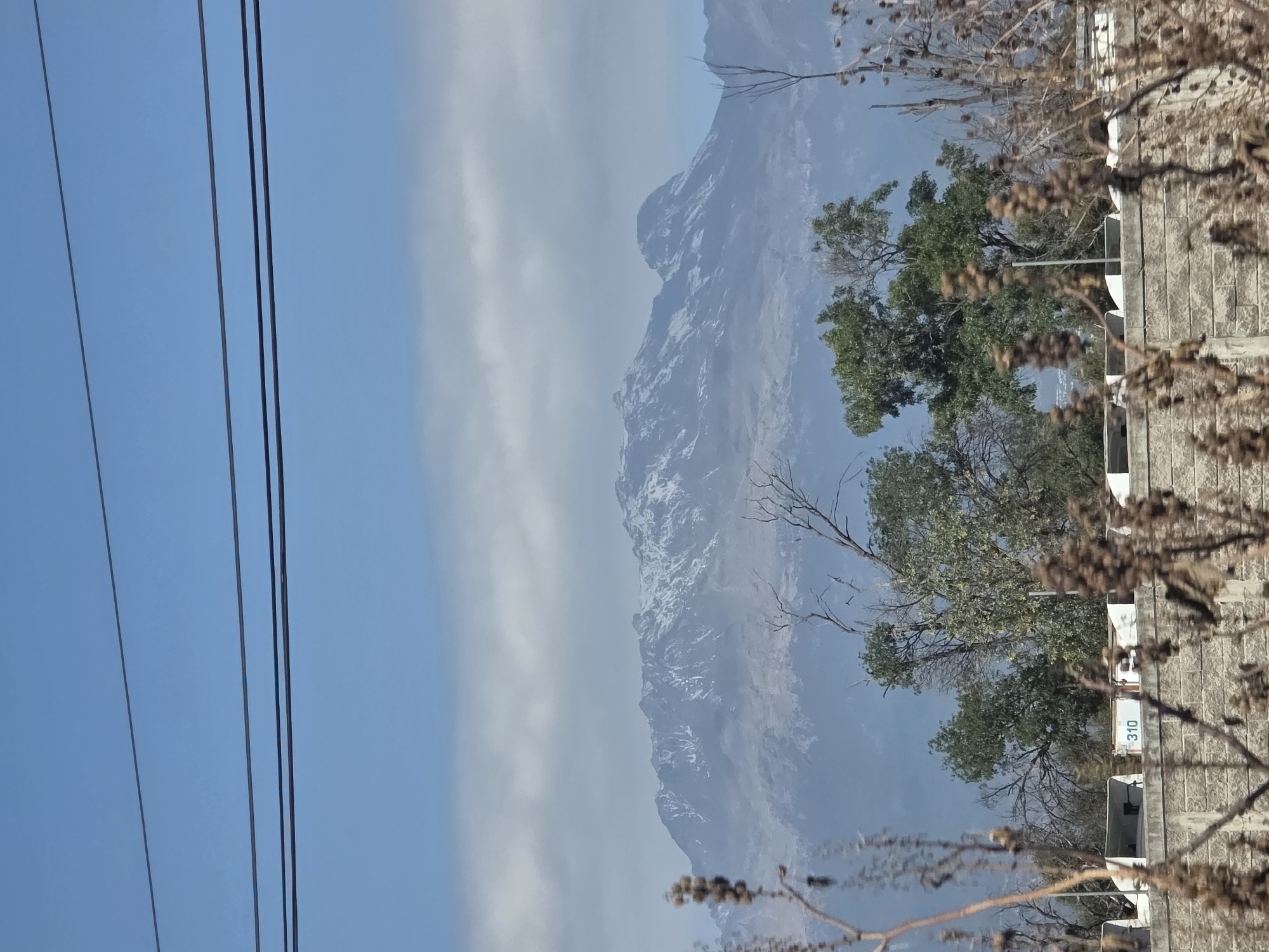 The image shows a mountainous landscape partially obscured by haze. The mountain range has snow-capped peaks. In the foreground, there are trees with green foliage and bare branches. Power lines stretch across the upper left portion of the frame. A building with the number "310" visible on its side is located on the right of the frame, partially obscured by branches. The location is Santa Ana Xalmimilulco, Mexico.