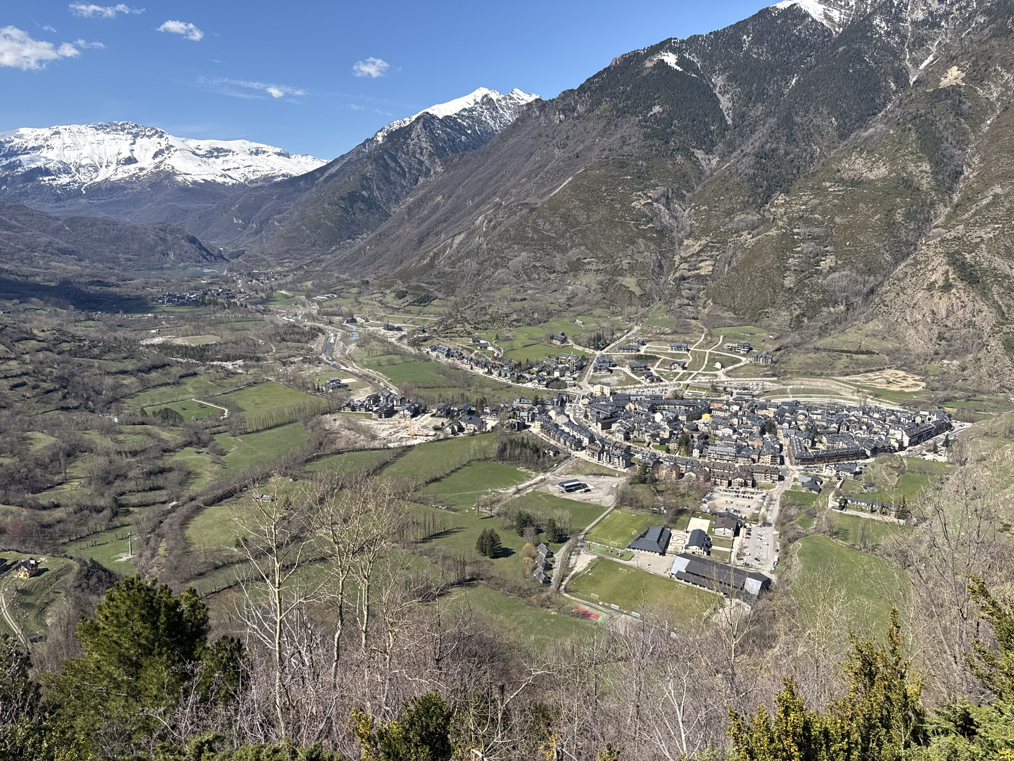 This is an aerial view of the town of Benasque, Spain, nestled in a valley surrounded by the Pyrenees Mountains. The foreground features trees and vegetation leading down to green fields, which then transition to the town itself. The town is densely packed with buildings, and roads crisscross the landscape. There are recreational fields and buildings. In the background, snow-capped mountains rise steeply, with a clear blue sky above.