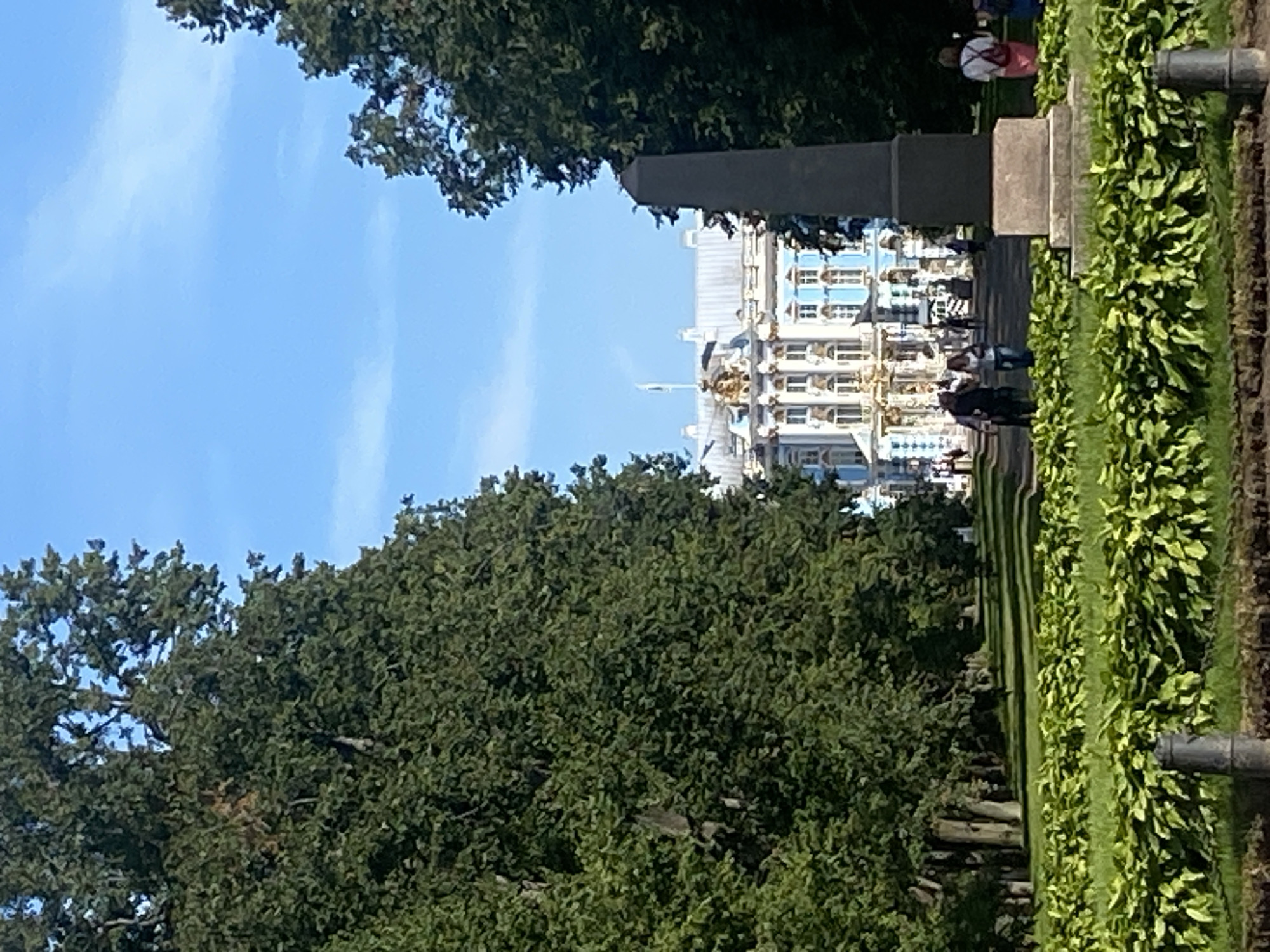 This image captures a view of the Catherine Palace in Pushkin, Russia, under a bright blue sky with scattered clouds. The palace, a grand structure with a light blue facade, white columns, and ornate gold detailing, is visible in the background. In the foreground, a pathway lined with tall, lush green trees leads towards the palace. A prominent, dark gray obelisk stands to the right of the pathway, in front of the palace. Several individuals are walking along the pathway, with some appearing to be tourists. The ground is covered with green grass and meticulously maintained hedges. The scene suggests a sunny day at a historical landmark, likely during a period of good weather for outdoor activities.