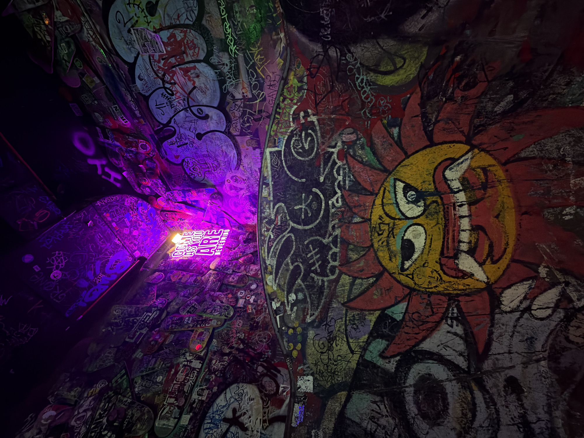 This image captures a vibrant and heavily graffitied indoor space, likely a tunnel or underpass, characteristic of urban art scenes. The scene is dominated by a myriad of graffiti tags, drawings, and stickers covering almost every visible surface, creating a dense, layered aesthetic.
On the right side, a prominent artwork depicts a stylized sun-like figure with a yellow, round face and large, white eyes featuring black pupils. Its mouth is open, revealing teeth, and it's surrounded by pointed, red rays, giving it an intense, almost angry or fierce expression. This central figure is a bold focal point amidst the surrounding visual chaos.
The space is illuminated by artificial lighting, with a strong purple hue emanating from the left, casting a dramatic glow on the surfaces and highlighting parts of the graffiti. This lighting creates a nocturnal or underground atmosphere. In the mid-ground on the left, a light source reveals what appears to be a neon sign or illuminated text, with parts of words like "DREAM" and "BE BRAVE" visible. Further to the left, affixed to the wall or ceiling, several skateboards are visible, also adorned with stickers and graffiti, suggesting a connection to skate culture.
Visible text includes numerous illegible tags and signatures. More distinctly, the word "SK8NG" is noticeable on the far left, "BRISANNA" on one of the skateboards, and possibly "GAVAS" within other tags. The overall impression is one of dynamic, unauthorized artistic expression in a public or semi-public urban environment, common in cities like Barcelona, Spain. No specific activity or event is taking place; the image is a static capture of the decorated environment.