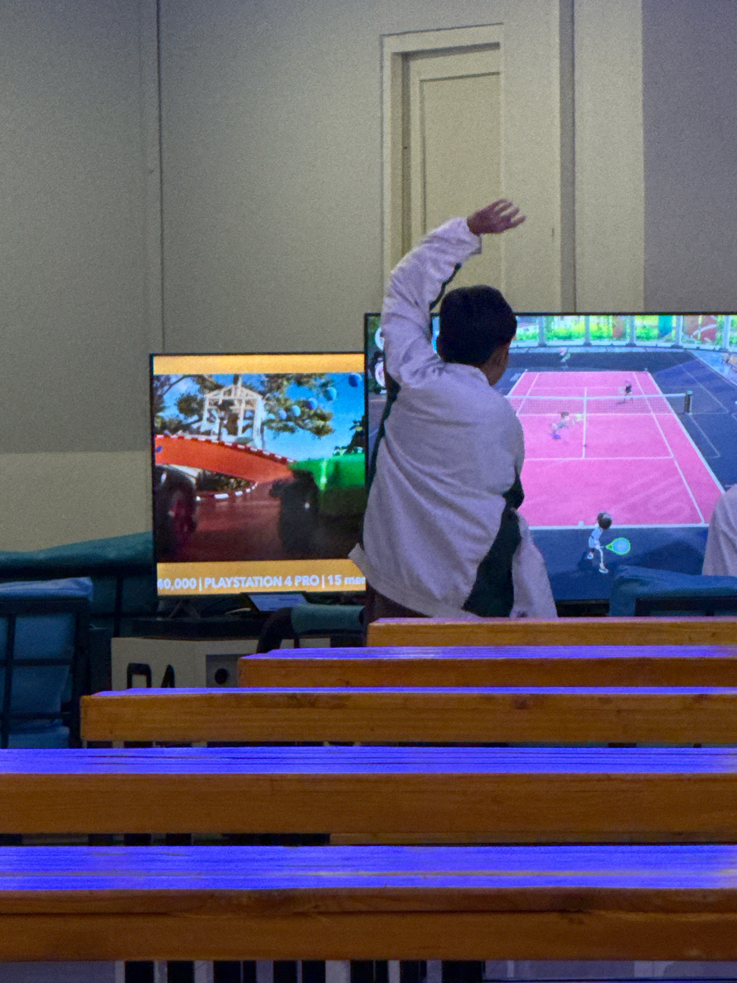 The image depicts a person from behind, wearing a white jacket over a dark undershirt, with their right arm raised. They are facing two television screens displaying video games. The left screen shows a scene with a red ramp and a structure resembling a gazebo, with text at the bottom indicating "40,000 | PLAYSTATION 4 PRO | 15 me". The right screen shows a tennis game with cartoonish figures on a pink court. The person appears to be playing or watching the games, as suggested by their posture. In the foreground, wooden benches with blue lighting are visible, indicating a seating area. The overall environment suggests an entertainment venue. There is a door frame visible in the background, and the wall appears to be a light grey color. Based on the provided crops and context, the location is tentatively identified as a PlayStation rental or gaming cafe in Surabaya, Indonesia, specifically in the Ruko Gateway Citraland area.