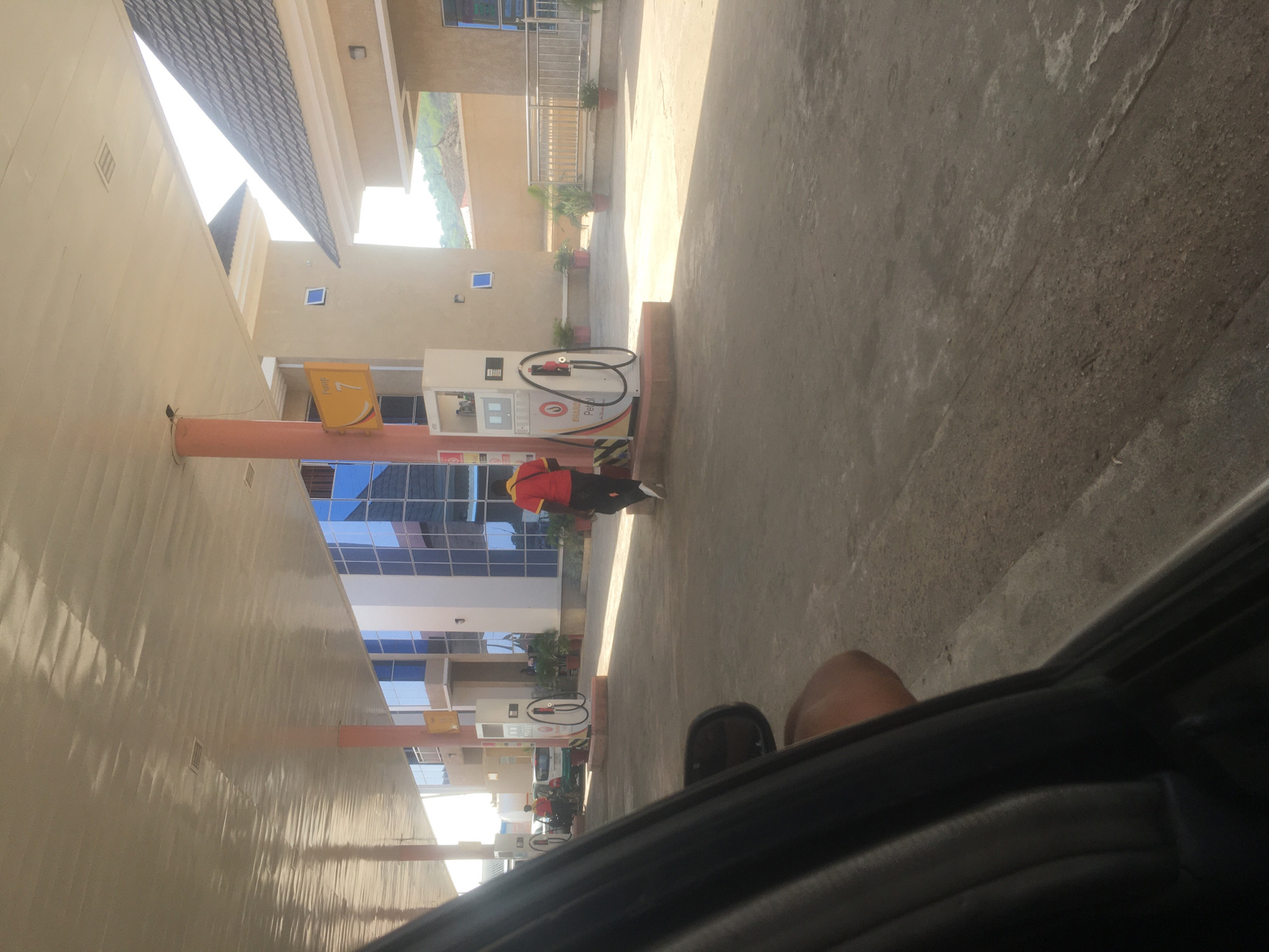 The image depicts a gas station in Jiru, Nigeria, taken from inside a car. A person in a red shirt is filling a vehicle with gas at a pump. The station has modern architecture, with a mirrored building and a covered area for the pumps. The ground is concrete. The image is oriented in portrait mode.