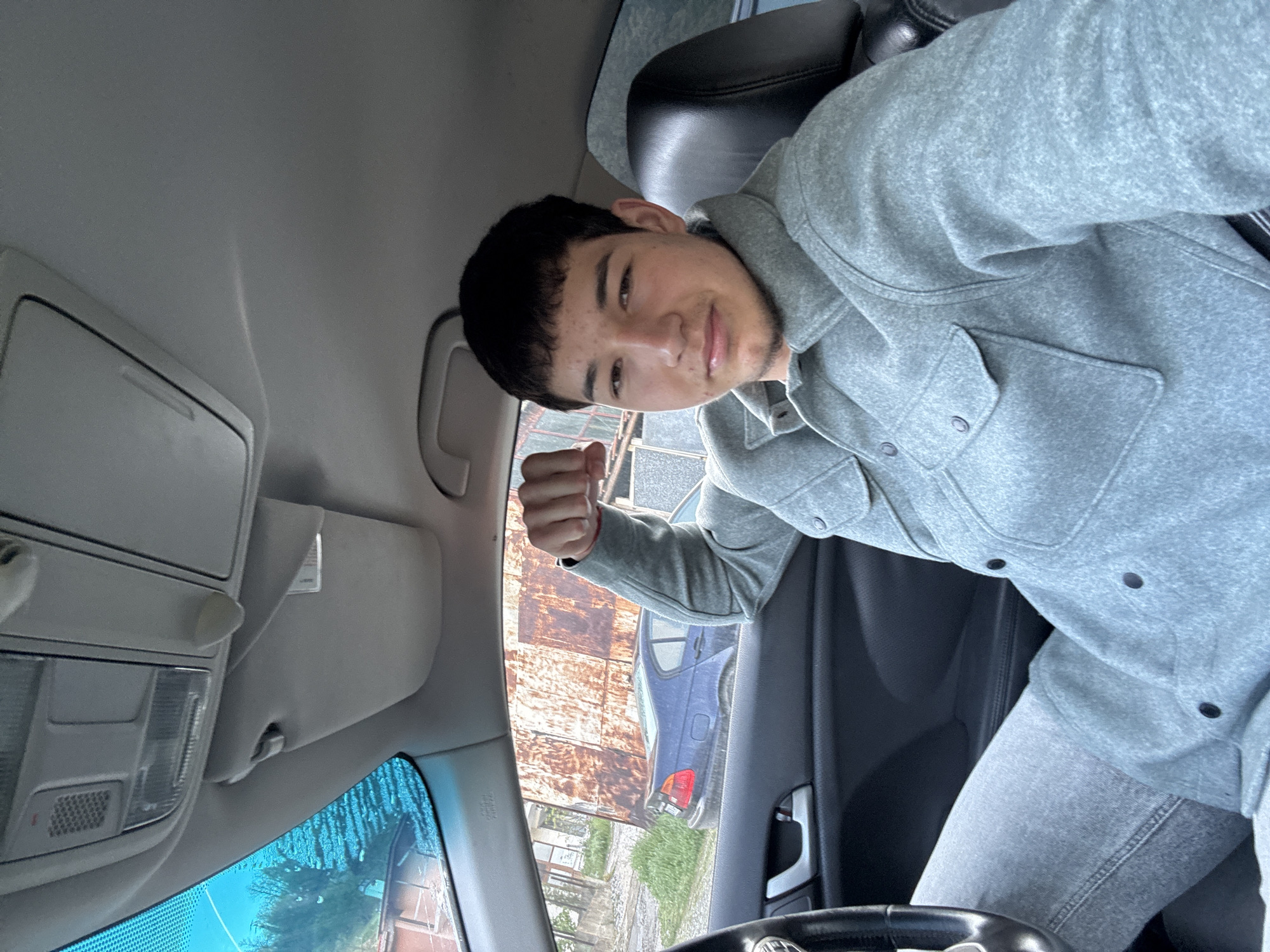 A young male sits in the driver's seat of a car. He is wearing a light gray button-up shirt and matching pants, and he is holding up a closed fist. The car's interior is gray and black. Through the window, a rusty corrugated metal fence is visible, as well as a blue car. The location is near Zlatograd, Bulgaria, based on the car's license plate visible through the front windshield.
